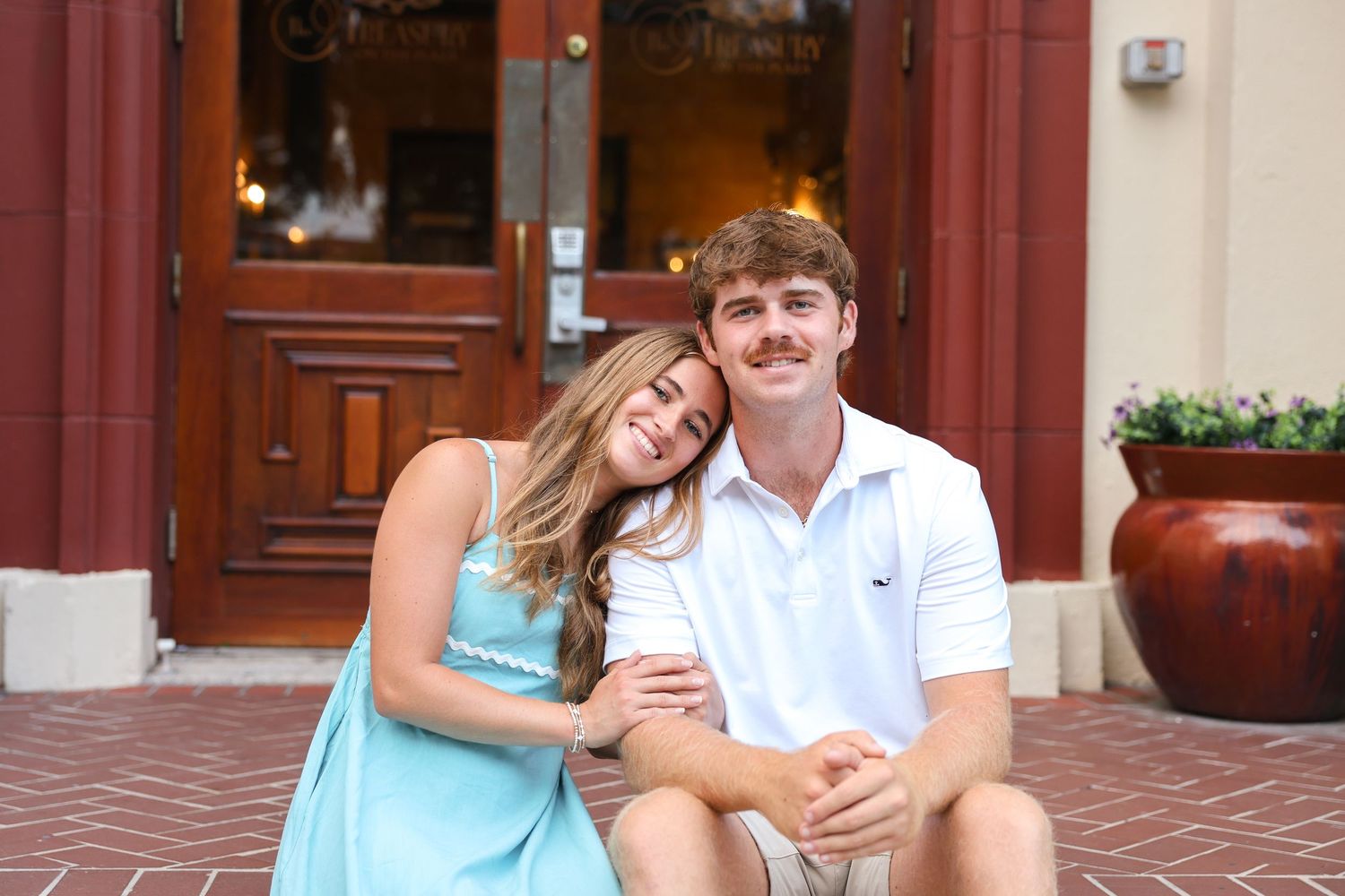 A young couple sits on brick steps, smiling, woman in blue dress leaning on man in white polo shirt outdoors.