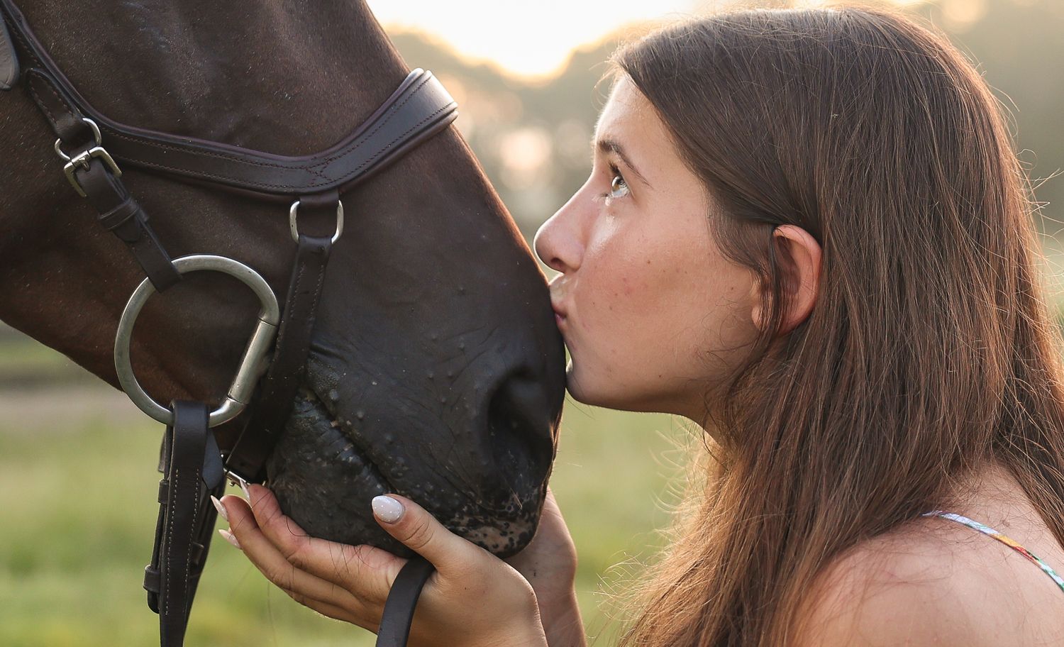 Young woman with long brown hair kissing a dark brown horse on the nose outdoors in natural light.
