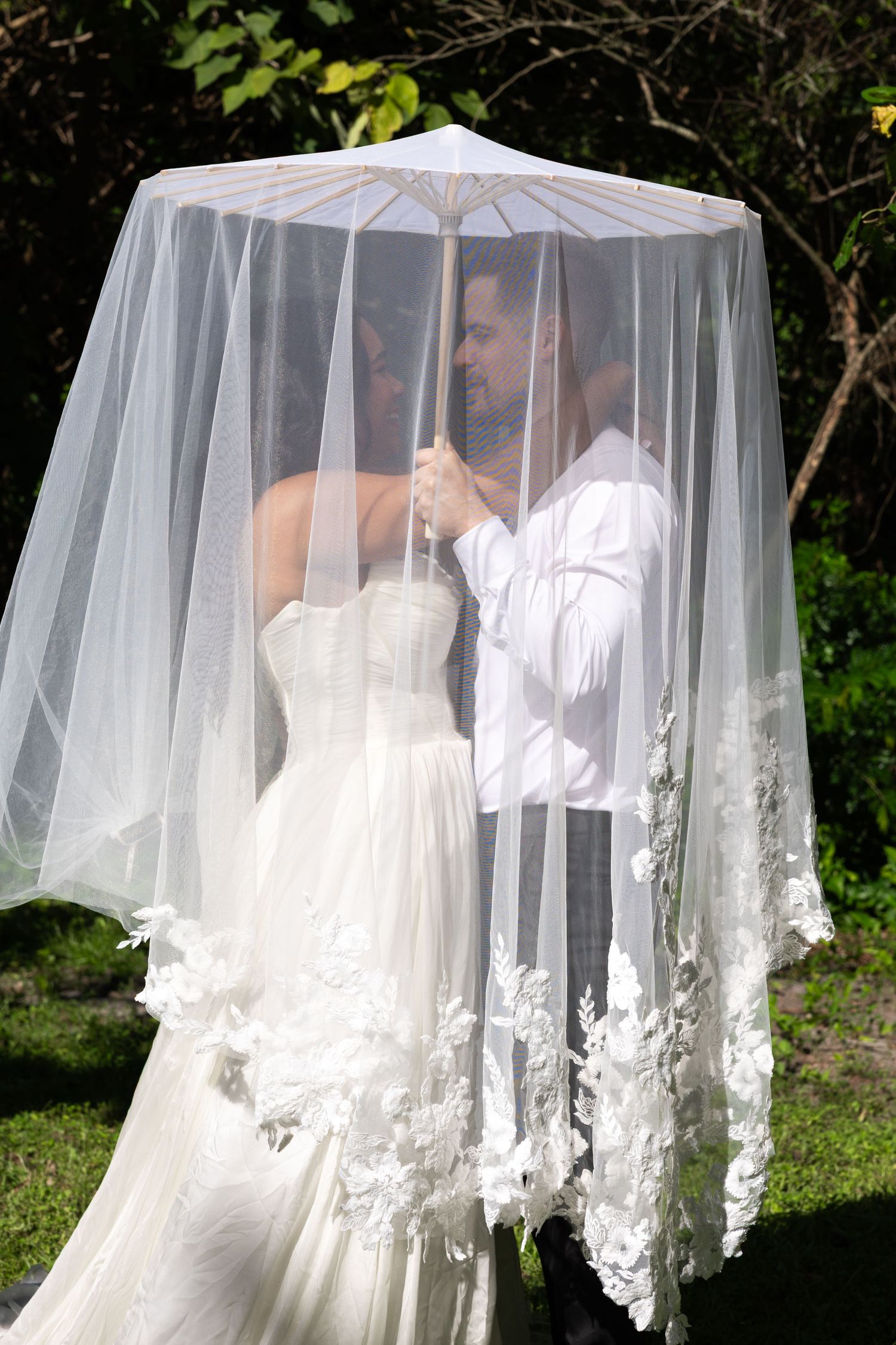 Bride and groom sharing a romantic moment under a white lace veil umbrella outdoors on their wedding day.