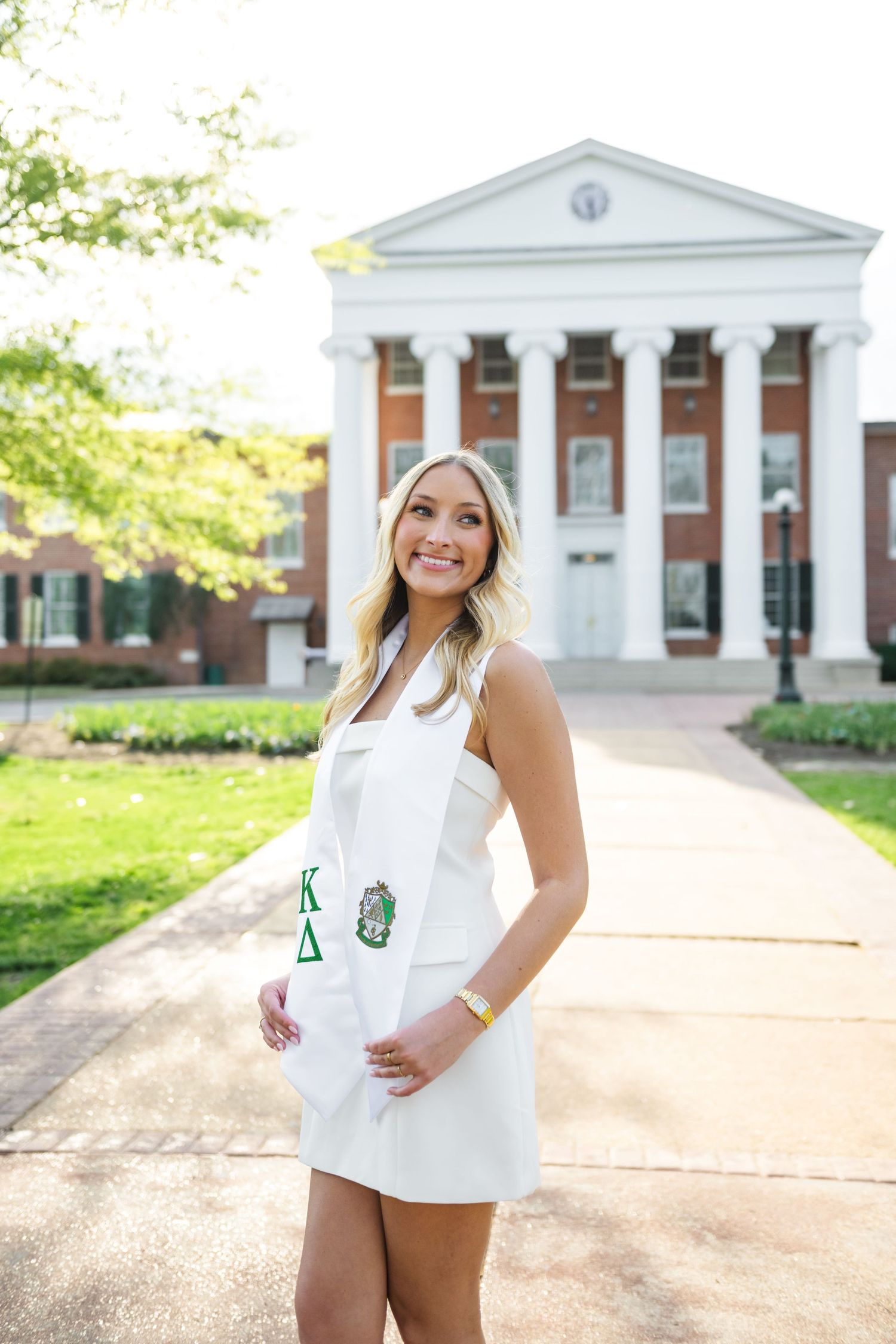 Smiling graduate in a white dress wears a Kappa Delta sorority stole and stands on a brick walkway before a columned campus building.