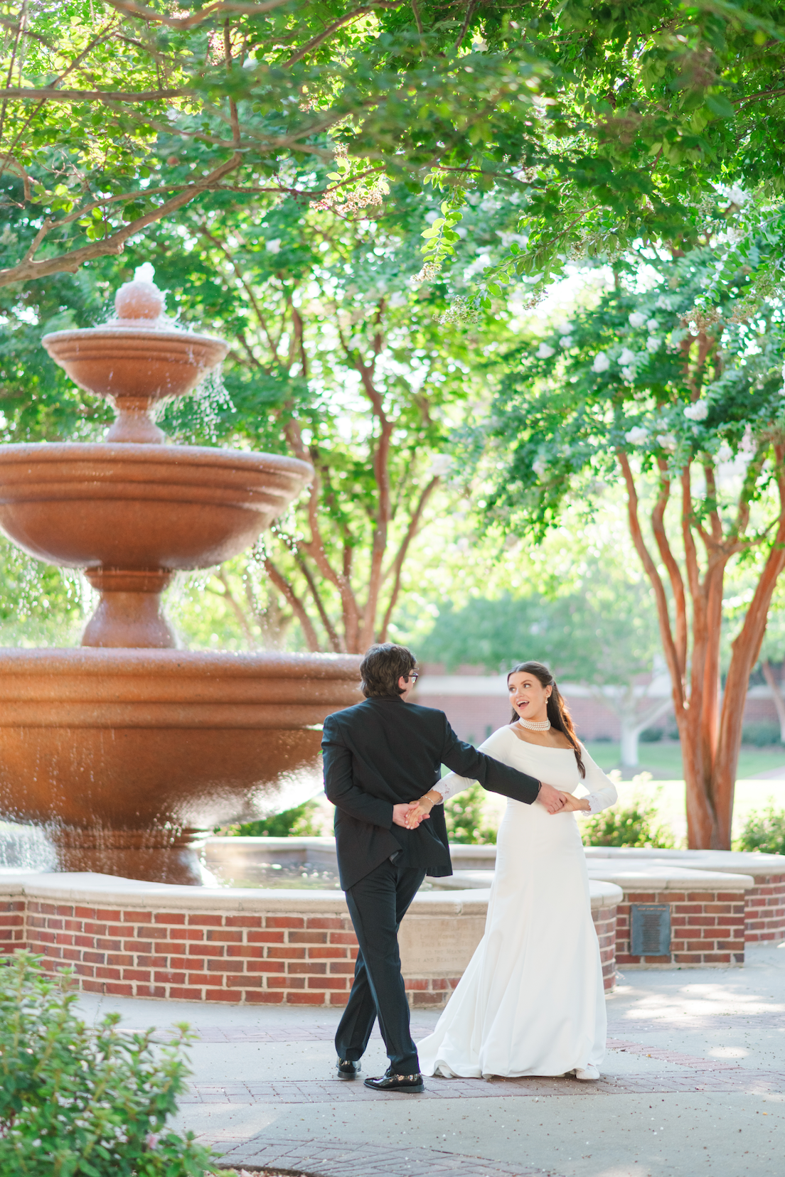 Bride in a white gown and groom in a black suit hold hands and dance near a tiered stone fountain beneath leafy trees.