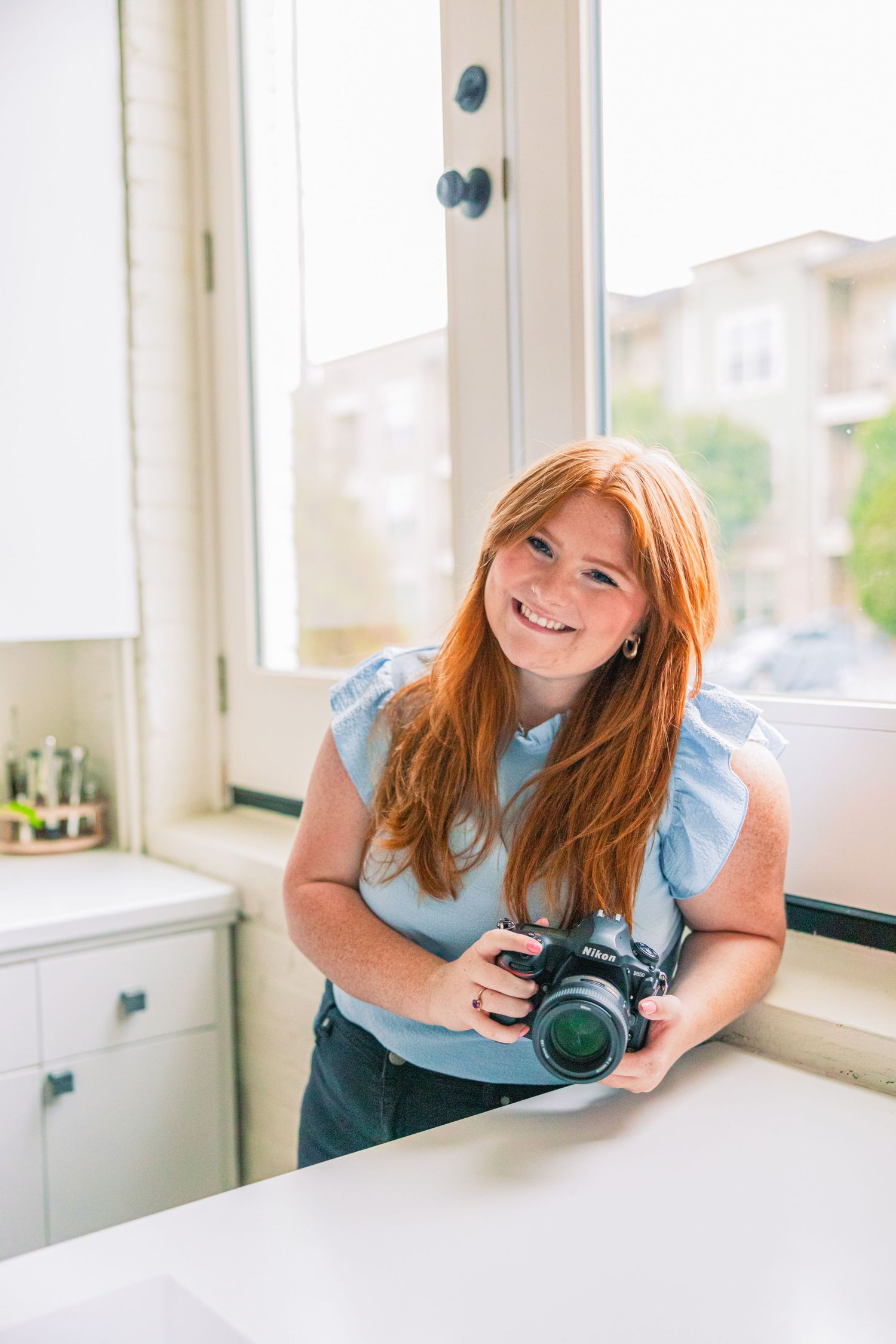 Smiling woman with long red hair in a light blue top holds a DSLR camera while leaning on a white counter beside a bright window.
