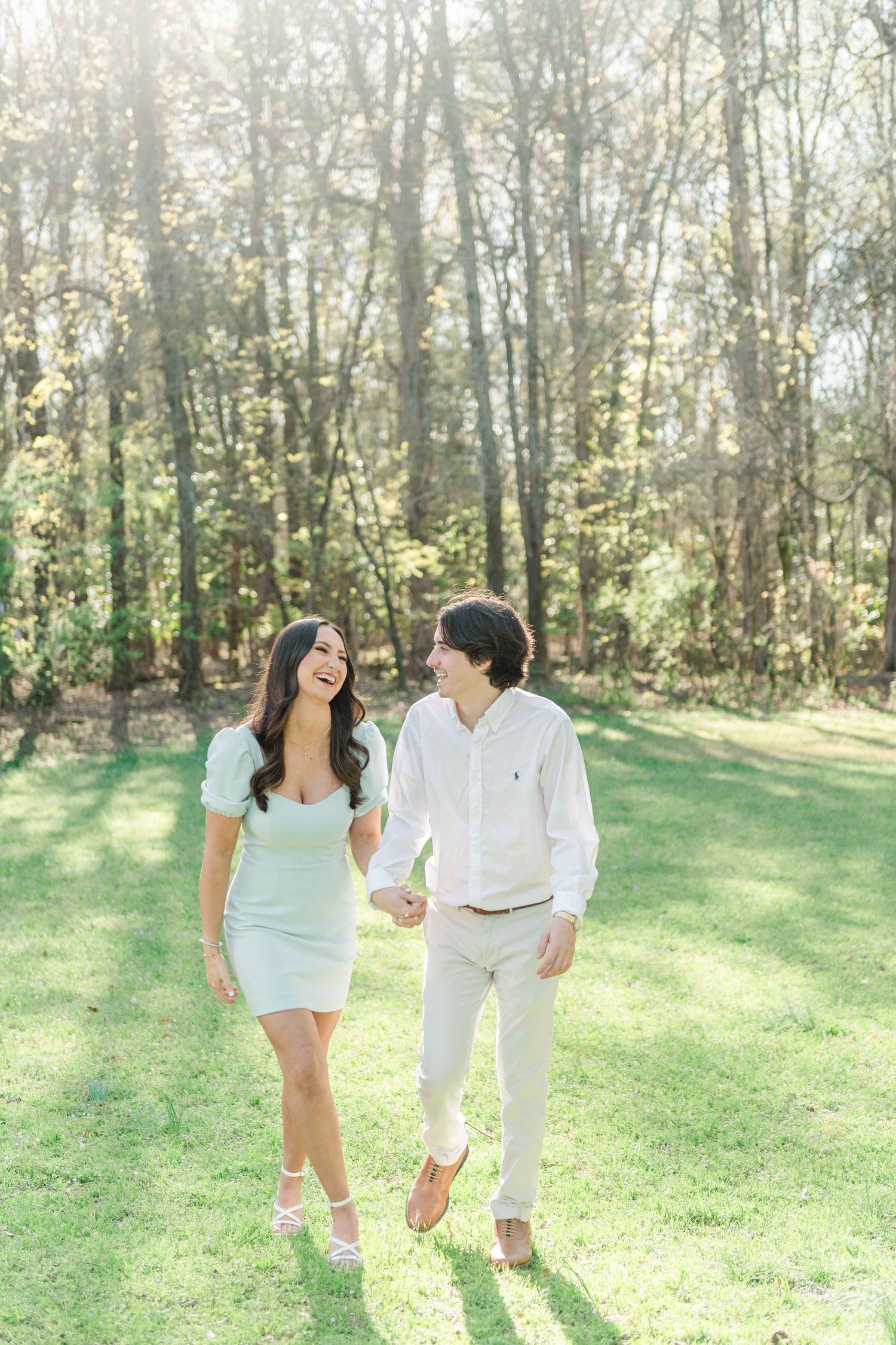 Smiling couple in light summer outfits hold hands and walk across a sunlit grassy clearing surrounded by tall trees.