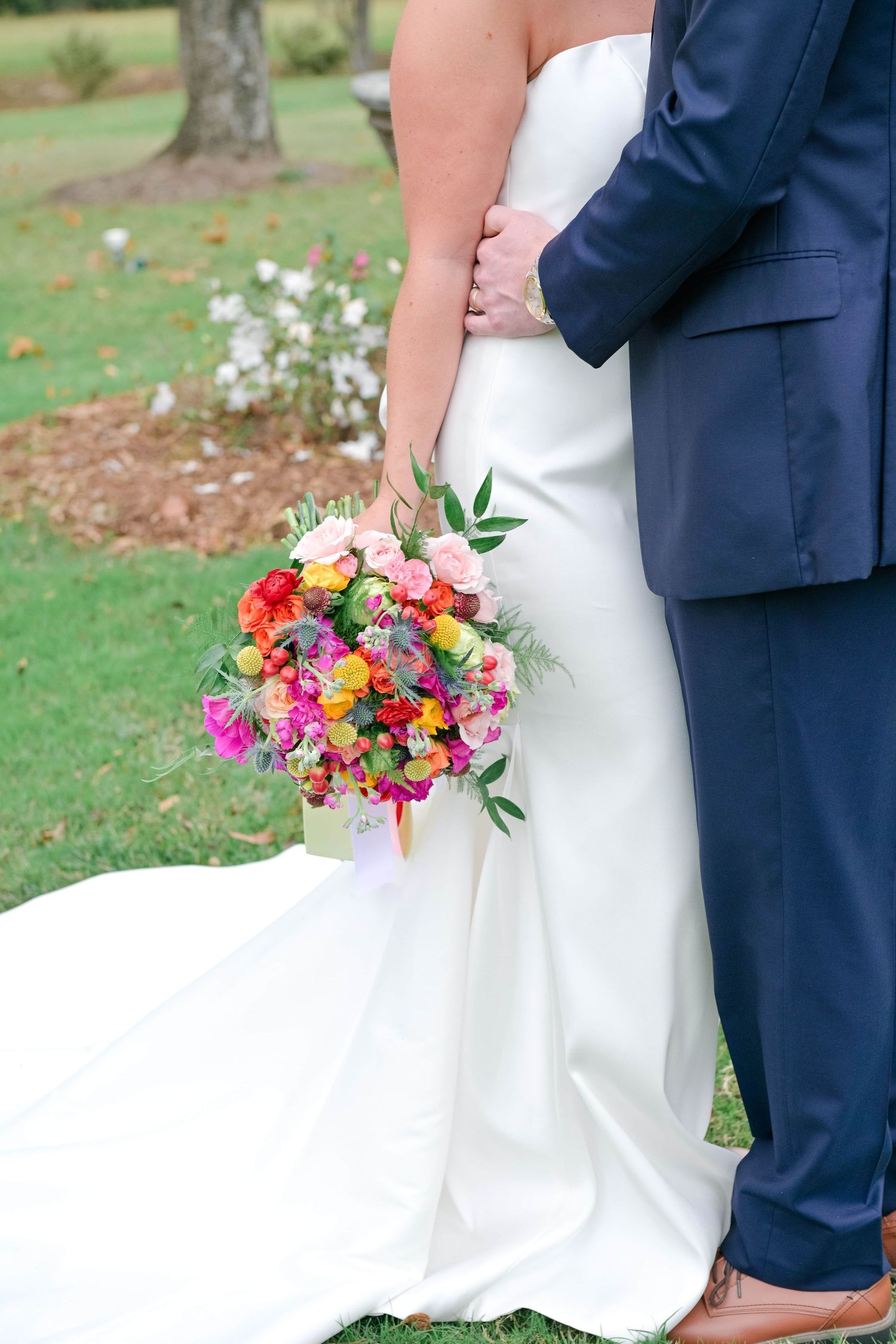 Bride in a white strapless gown holds a colorful bouquet of pink, orange and yellow flowers beside a groom in a navy suit on a lawn.