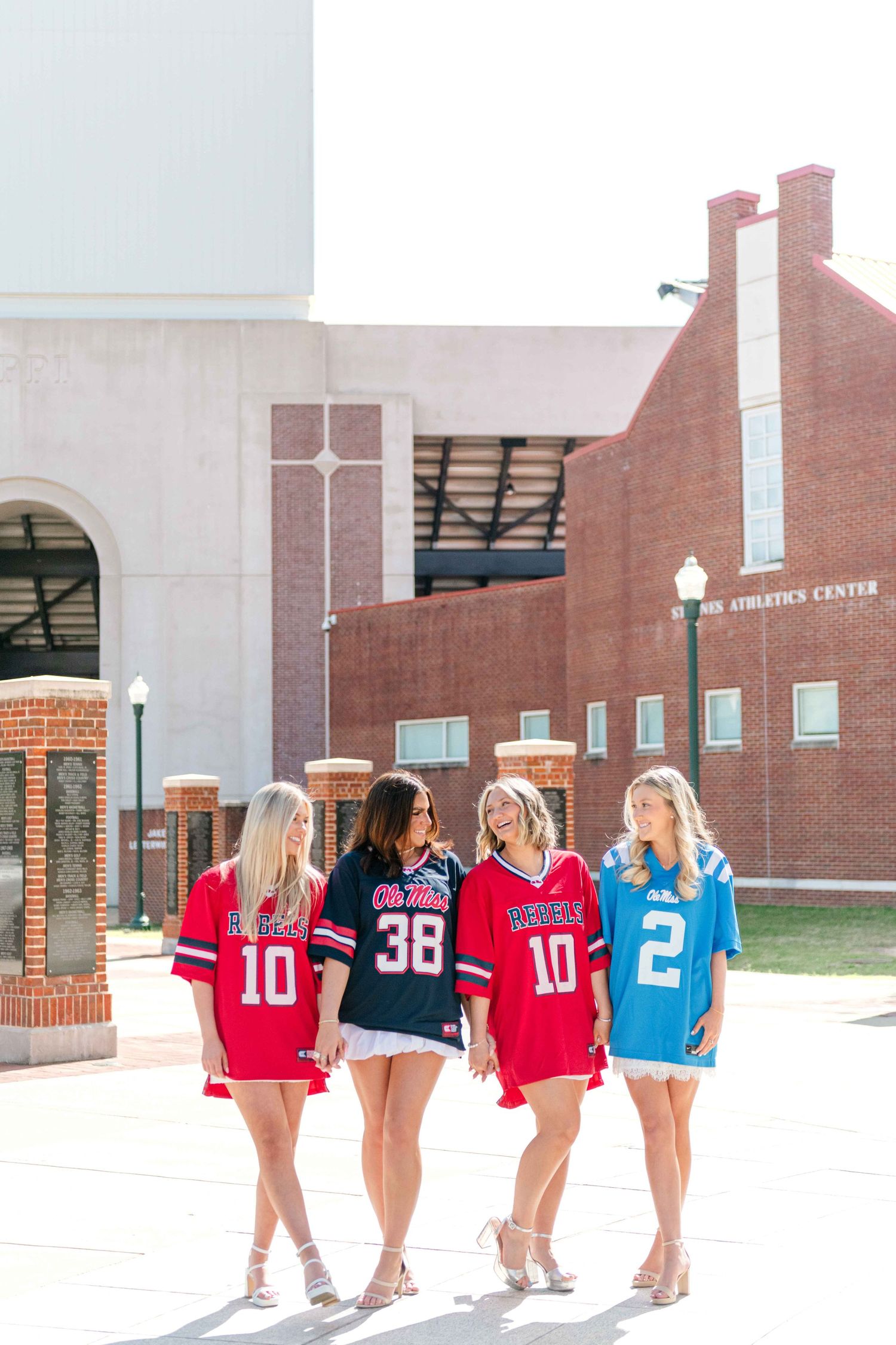 Four young women in red, navy, and light blue football jerseys walk arm in arm in front of a brick athletics center building.