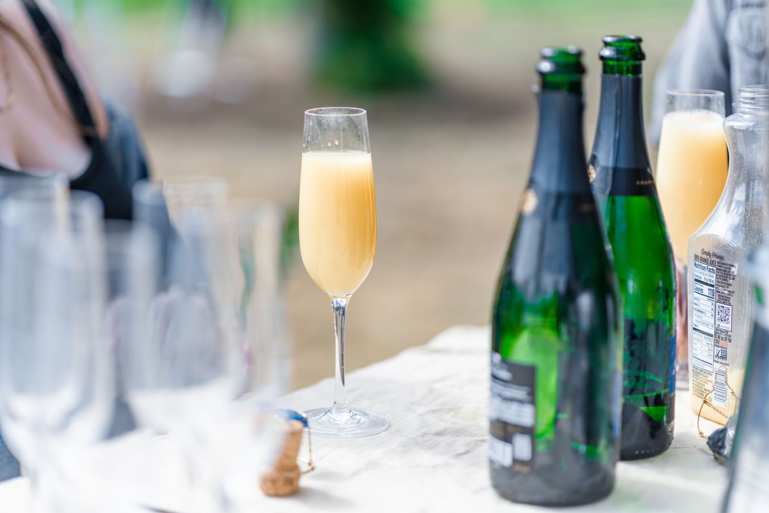 Champagne flute filled with a peach-colored mimosa sits on a table beside green sparkling wine bottles and empty glasses outdoors.