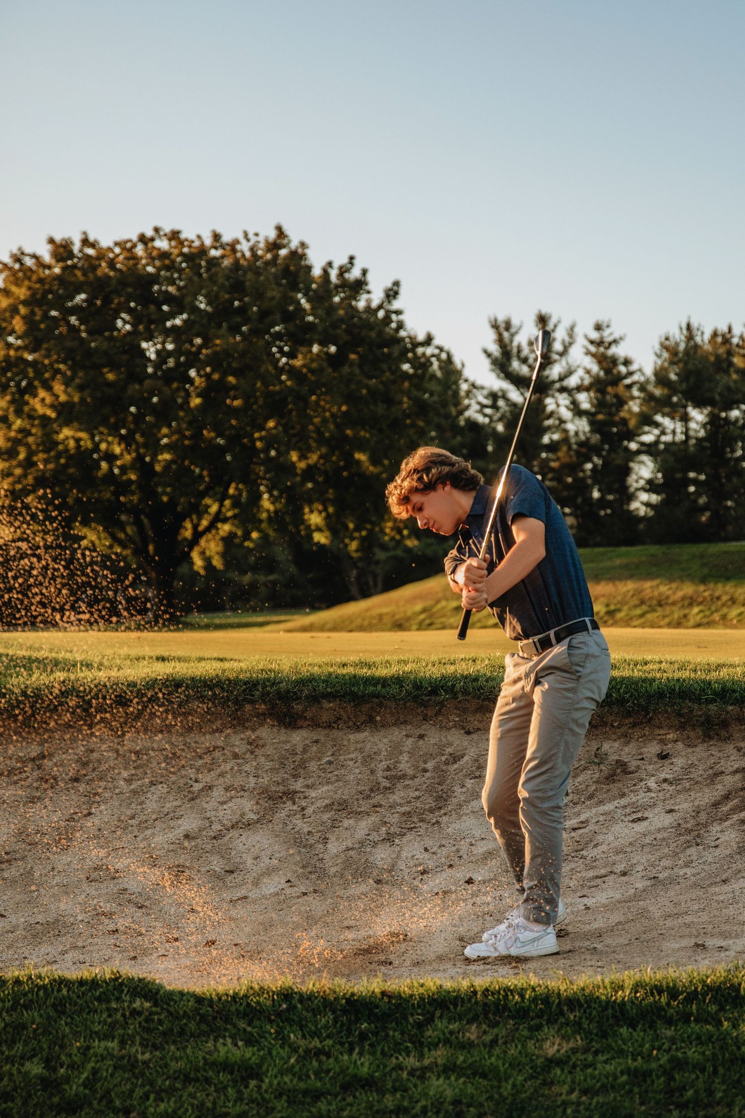 Young golfer executing a sand bunker shot on a sunny golf course, with sand flying and lush green trees in the background.