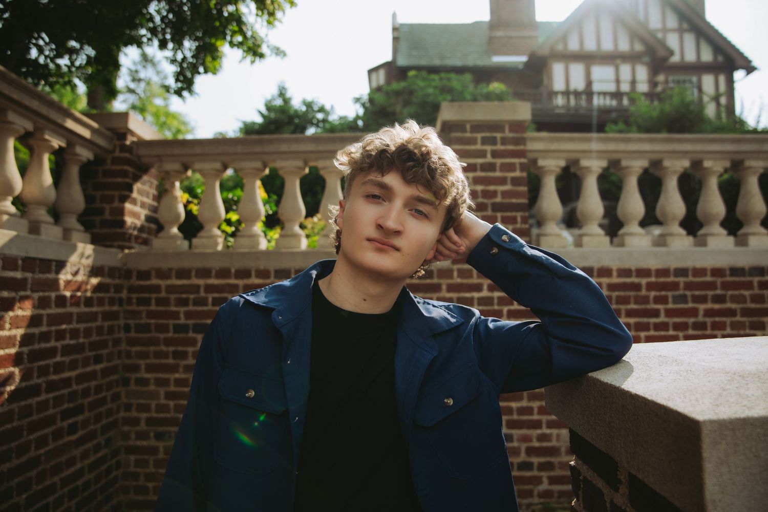 Young man with curly hair in blue jacket poses outdoors near brick wall and stone balustrade.
