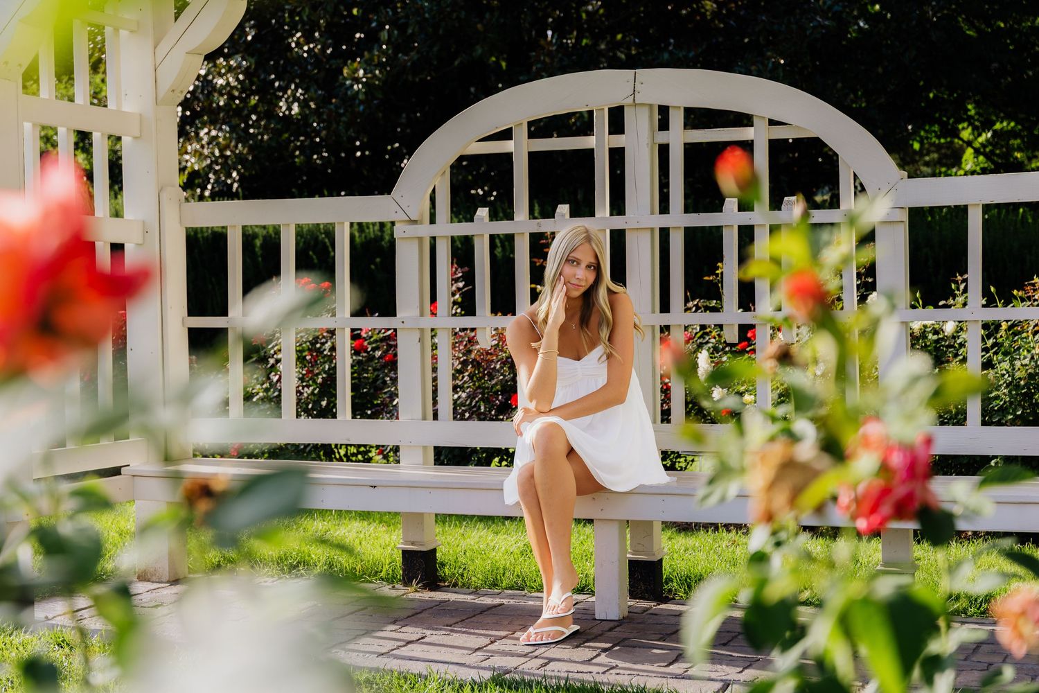 Young woman in white dress sitting on a bench in a rose garden gazebo on a sunny day.