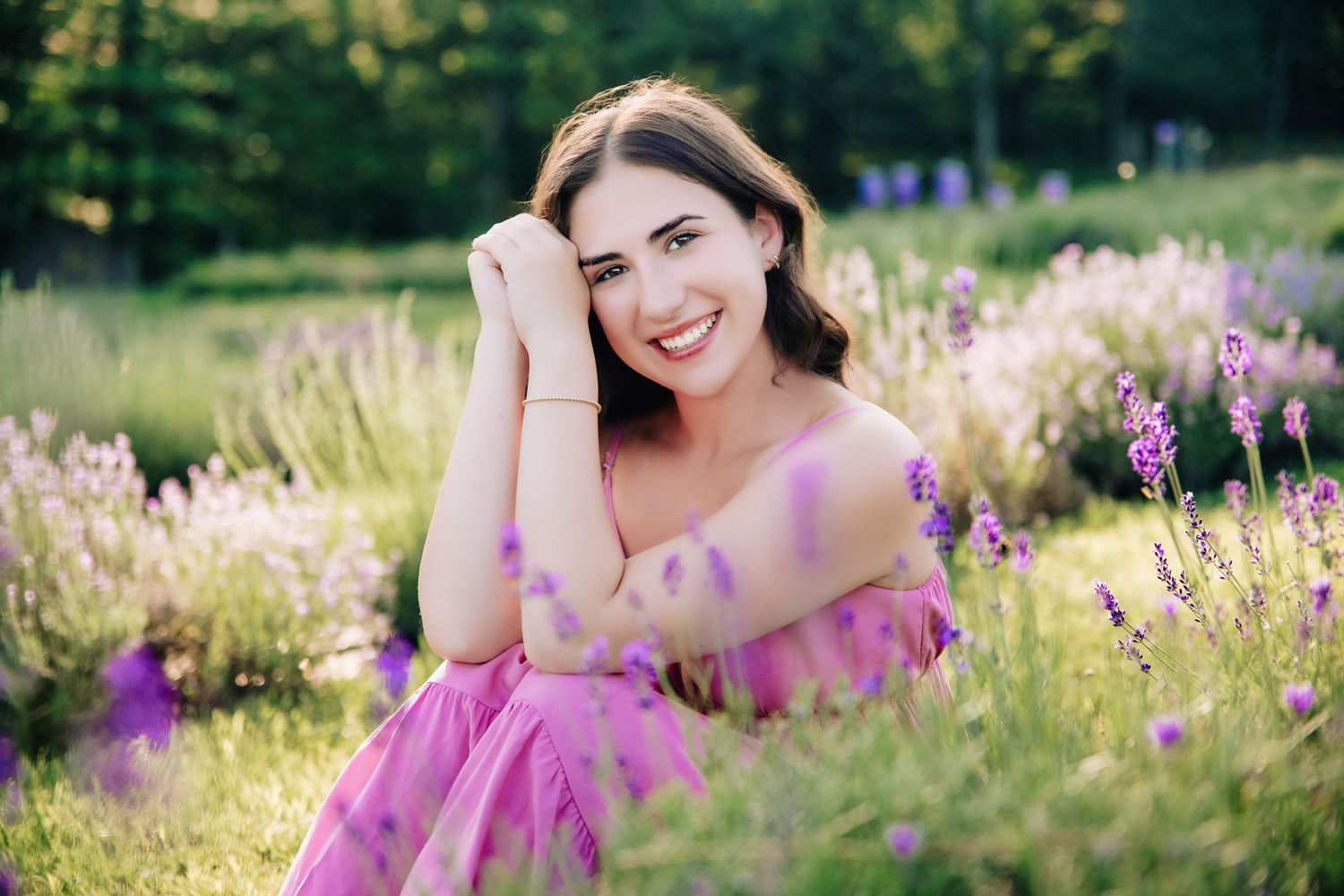 A smiling young woman in a pink off-shoulder dress sits among blooming lavender fields in soft golden sunlight.
