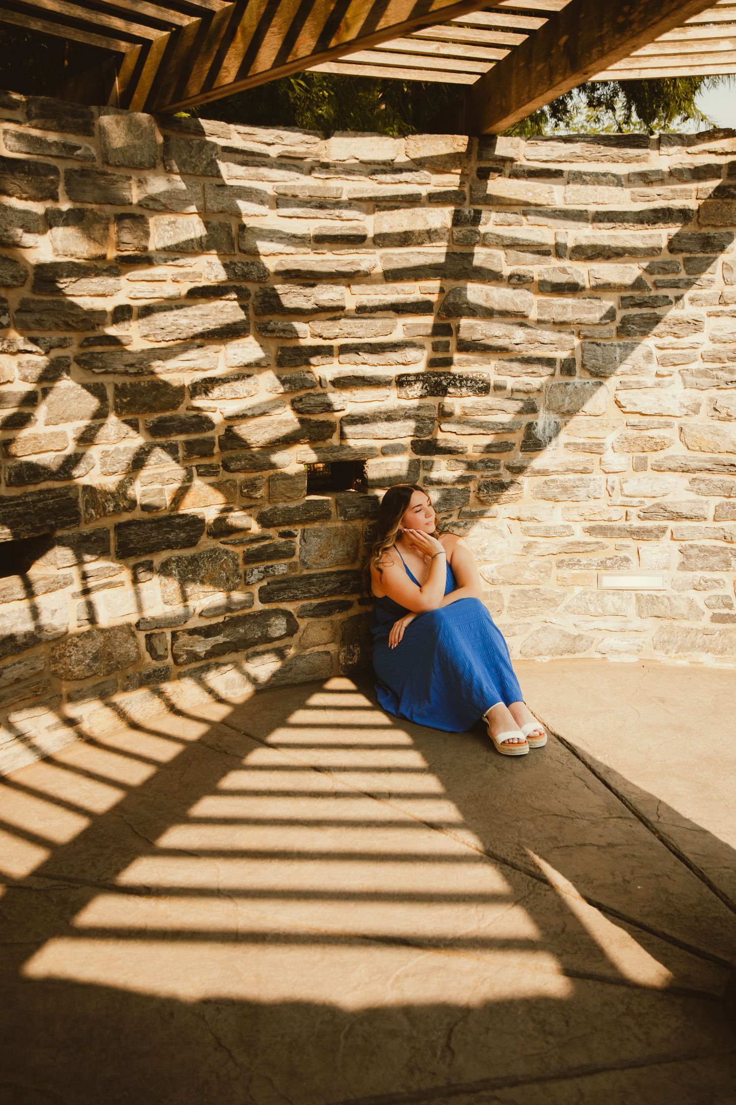 Woman in blue skirt sitting against stone wall under pergola with dramatic shadow patterns.