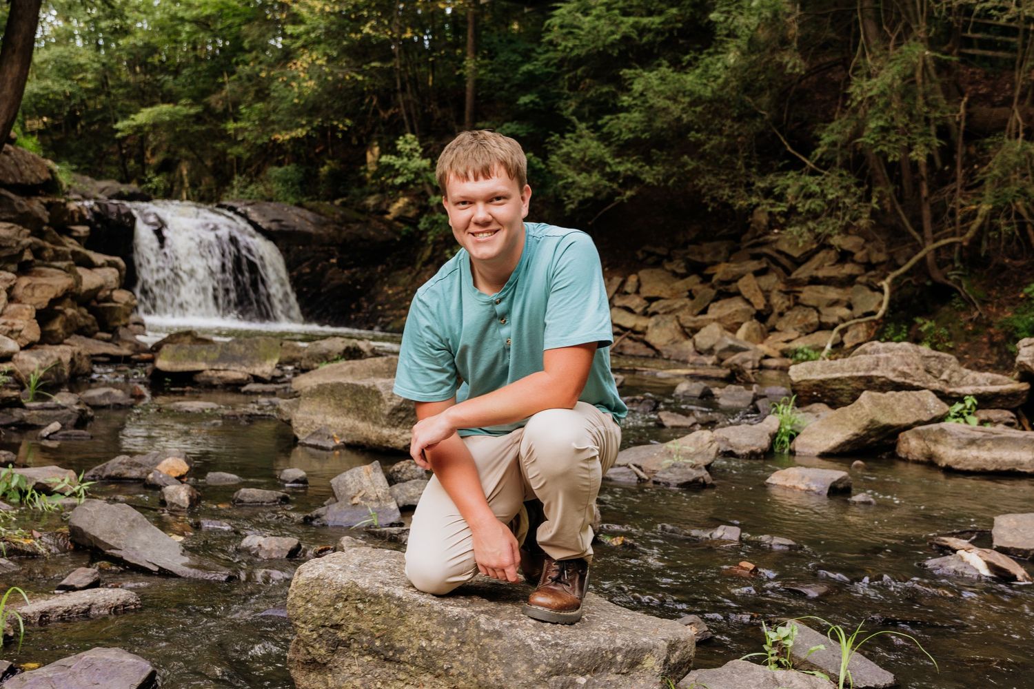 Young man in teal shirt crouching on rocks beside a serene woodland waterfall in a lush green forest.
