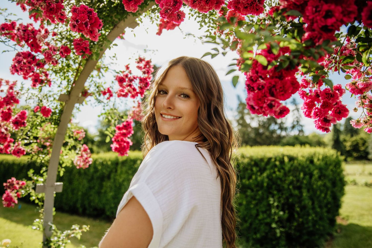Young woman in white top smiles over shoulder under vibrant red rose arch in sunny garden.