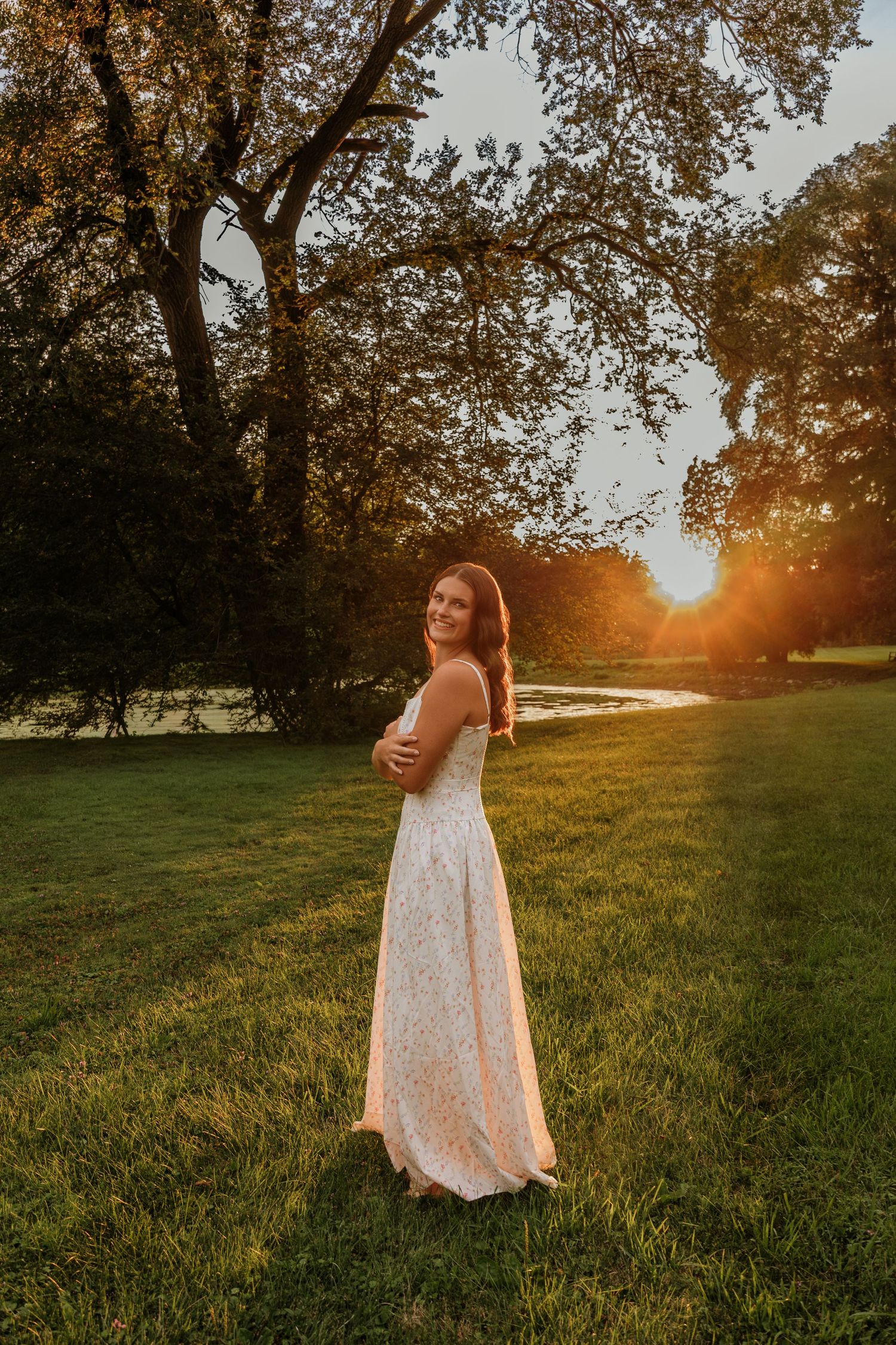Woman in white lace gown standing on green grass at golden sunset with trees and river in background.
