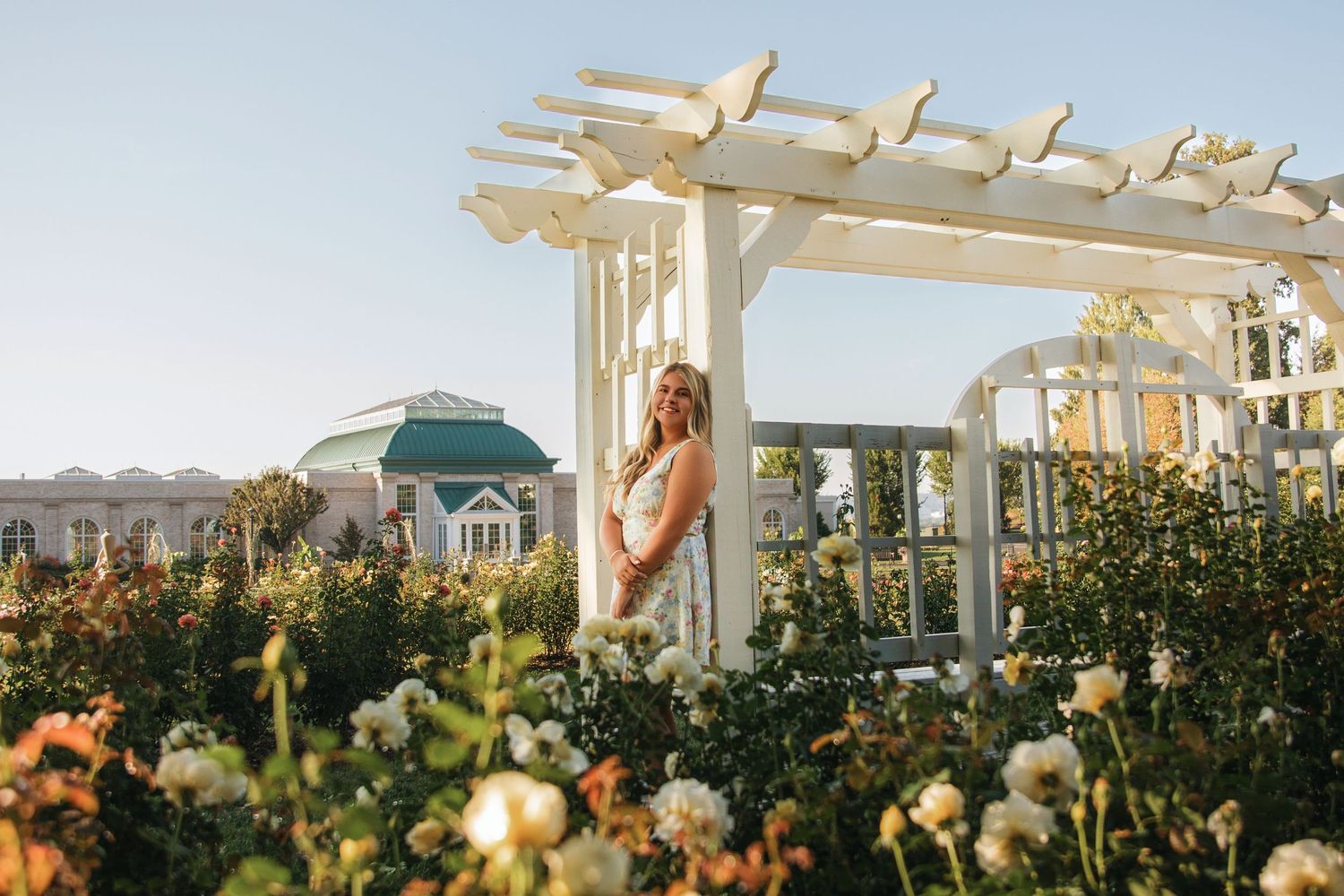 Woman in white dress standing under a white pergola surrounded by blooming roses in a garden.