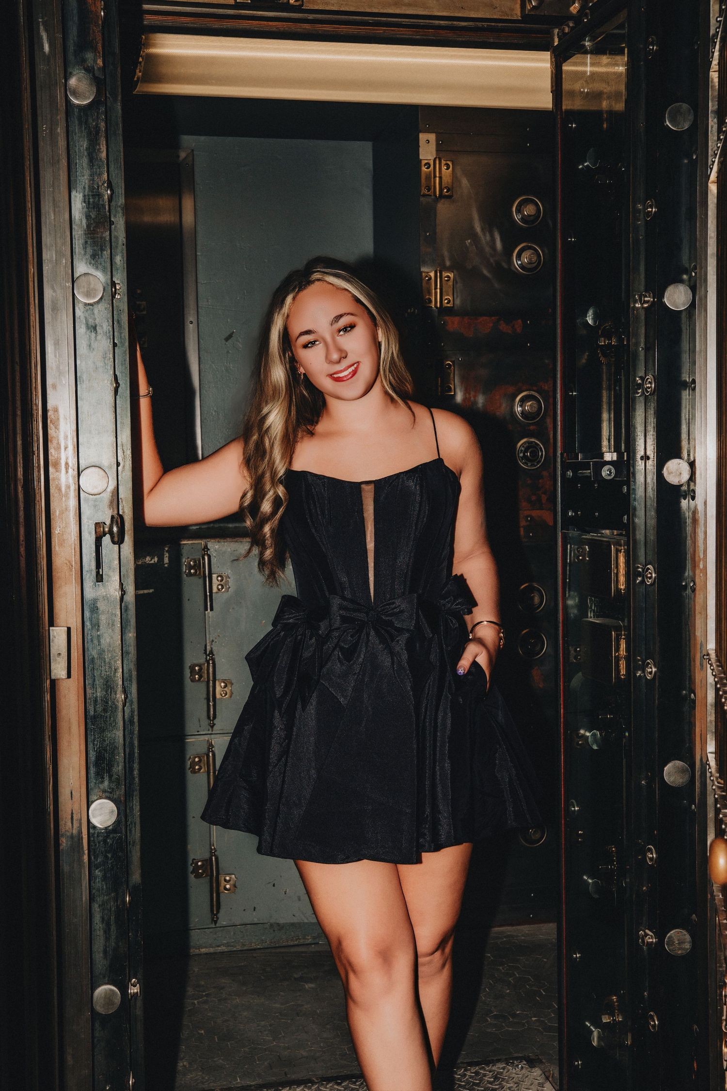 Young woman in black mini dress posing in vintage bank vault doorway, smiling confidently.
