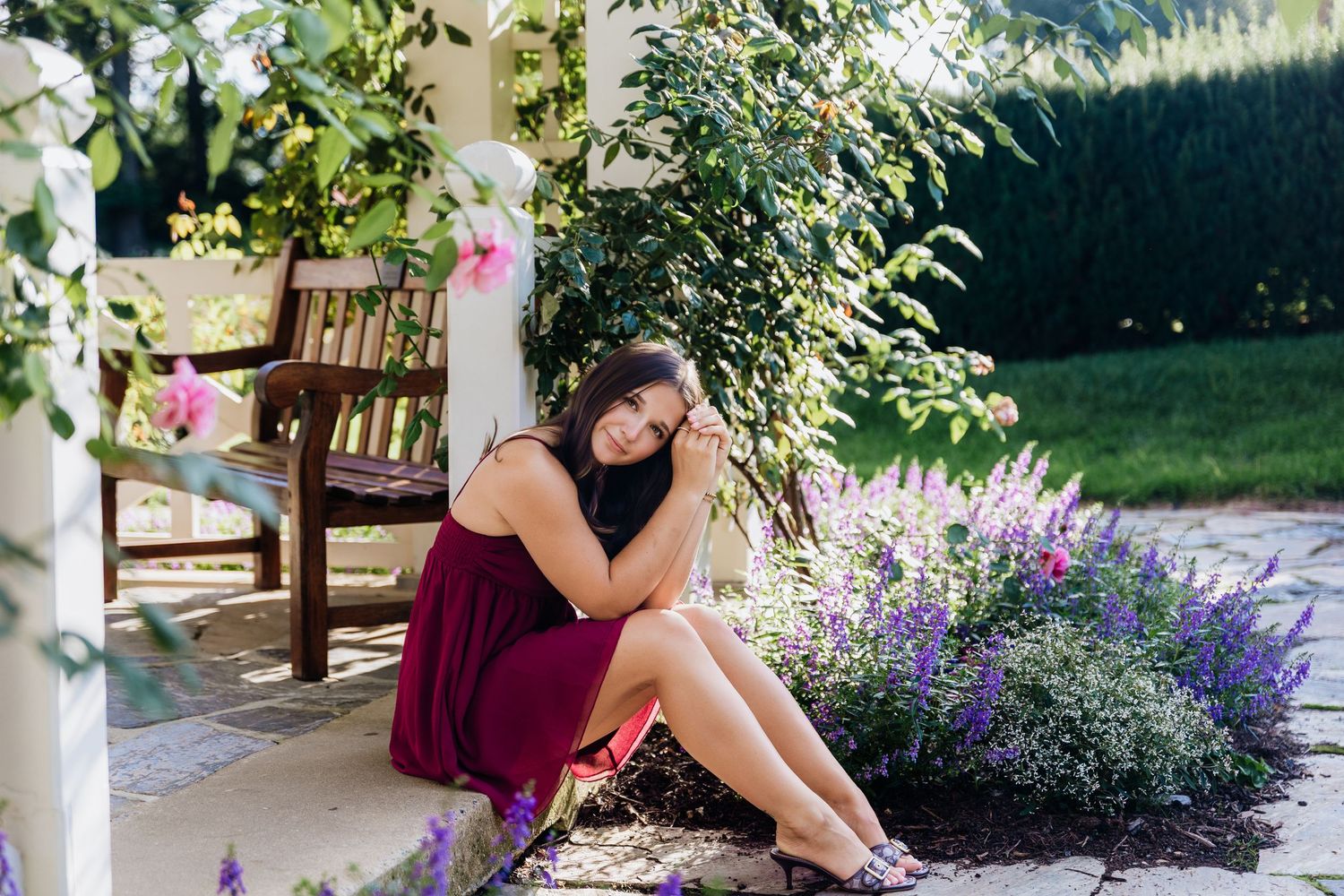 Young woman in burgundy dress sitting by garden flowers near a wooden bench on a sunny day.