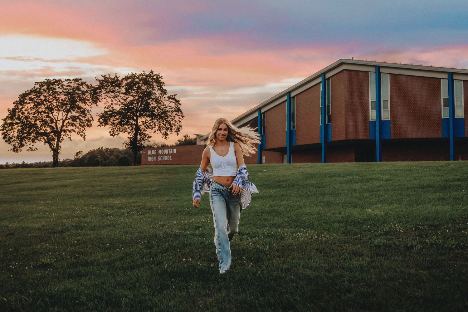 A young blonde woman walks across a green lawn at sunset, wearing jeans and a white top, with a colorful sky behind her.