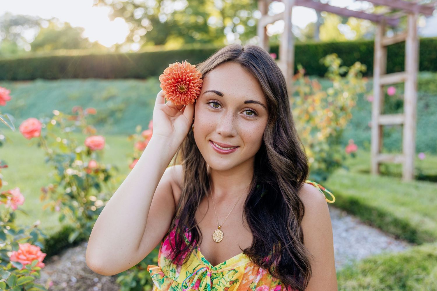 Young woman holding an orange dahlia flower in a lush rose garden wearing a colorful floral dress.