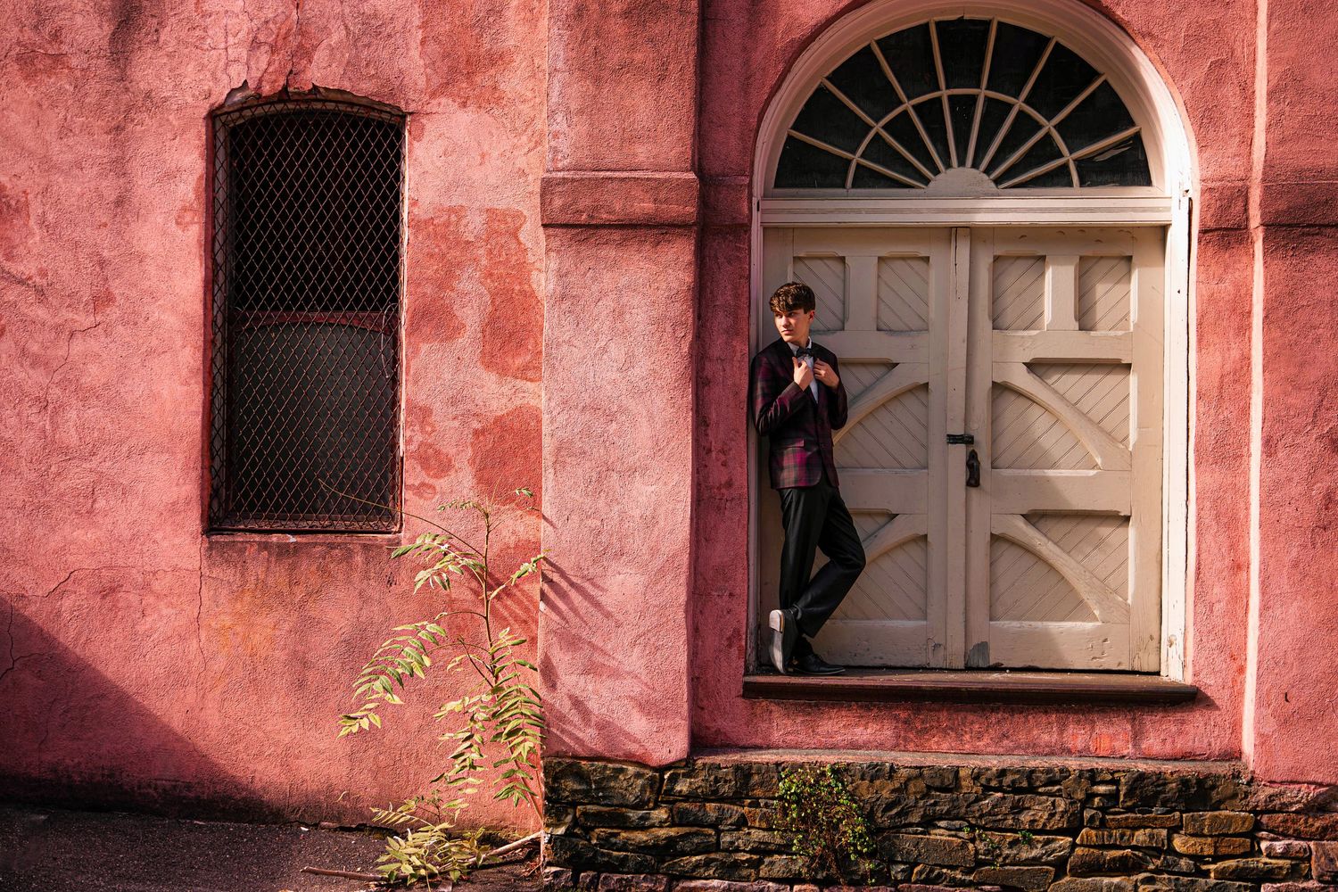 Person leaning against a white arched door on a pink building wall with a barred window nearby.
