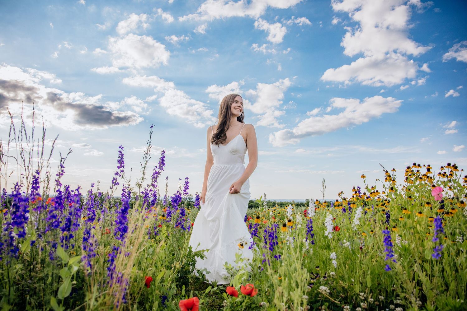 A bride in a white dress stands among vibrant wildflowers under a blue sky with fluffy clouds.