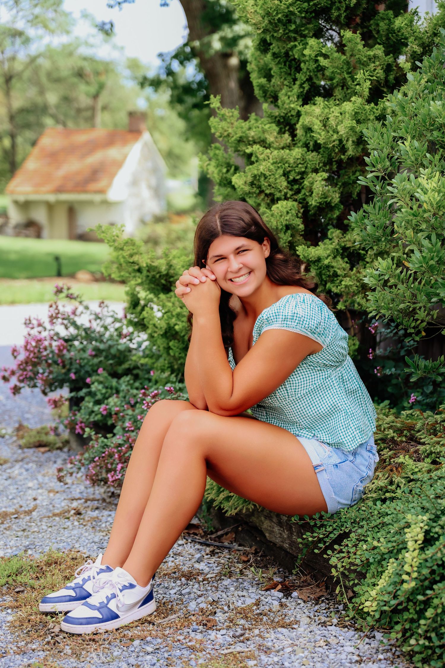 Smiling teenage girl sitting outdoors by green shrubs in denim shorts and blue sneakers with a cottage in the background.