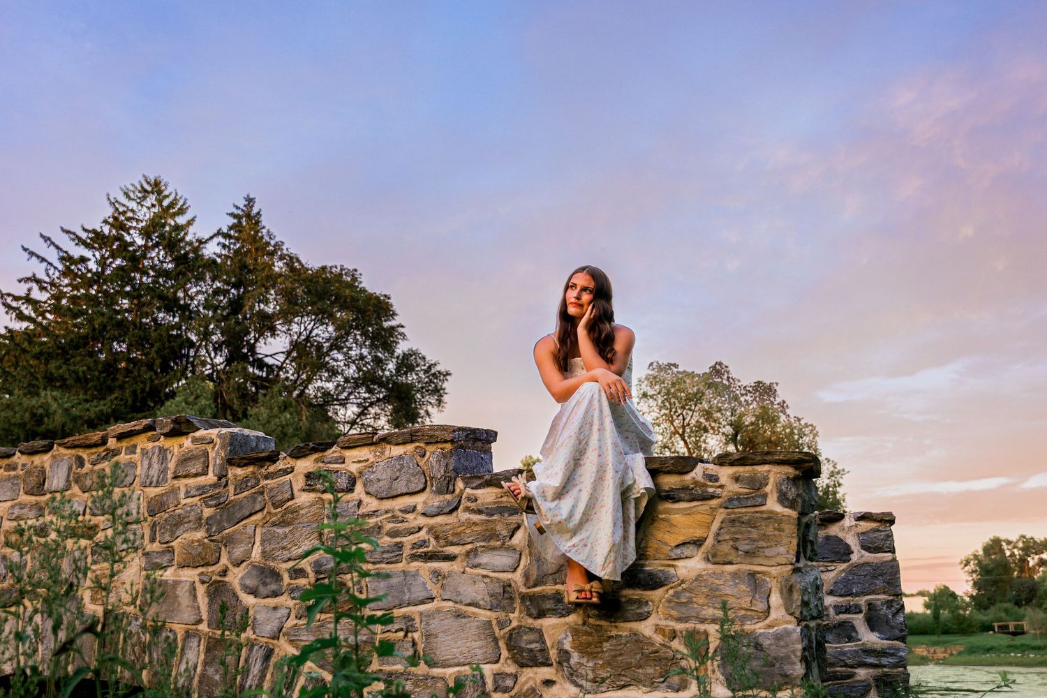 Young woman in white dress sitting on rustic stone wall at sunset with trees and colorful sky behind her.