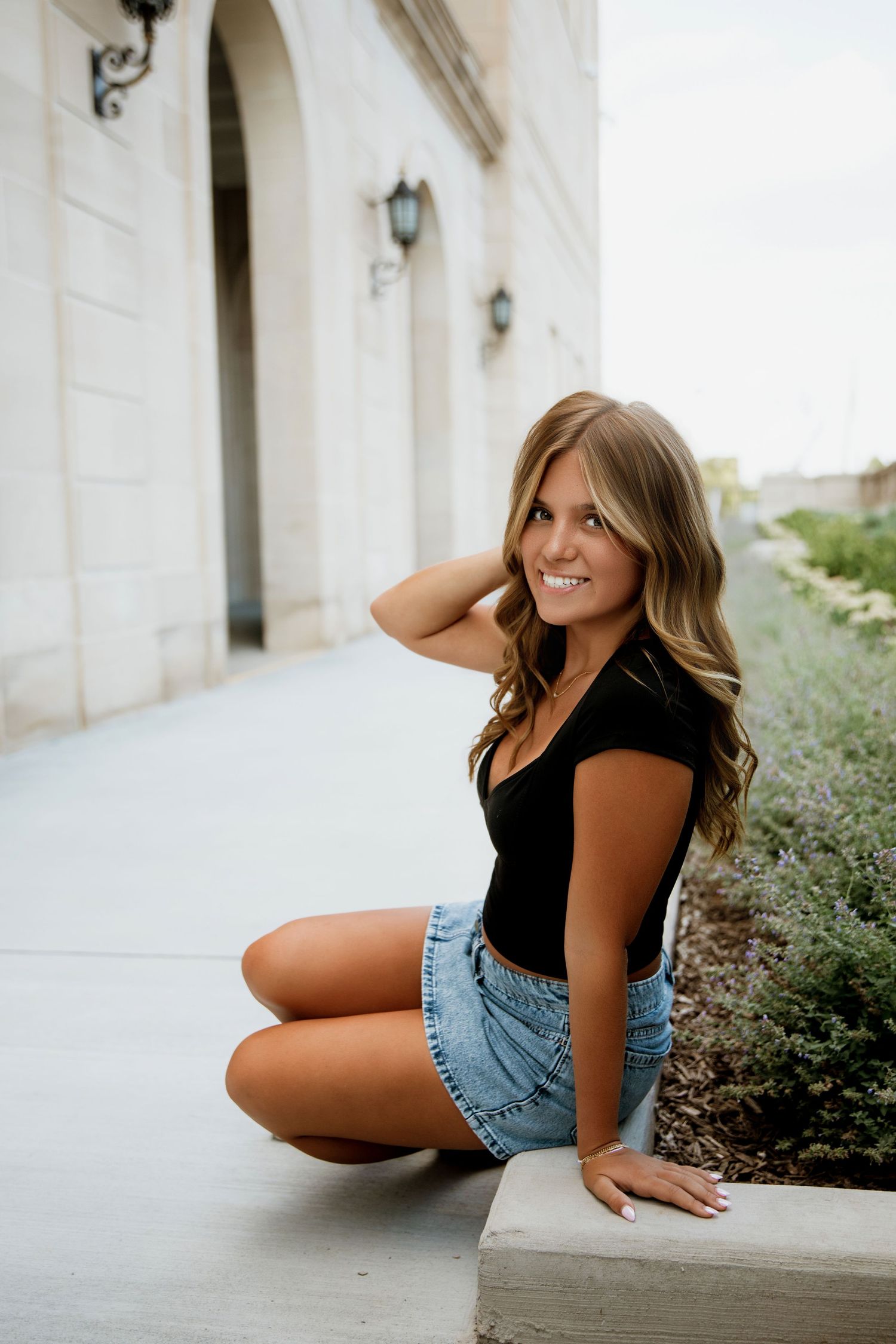 Young woman with long wavy hair sits outdoors smiling, wearing black top and denim mini skirt beside a stone building.