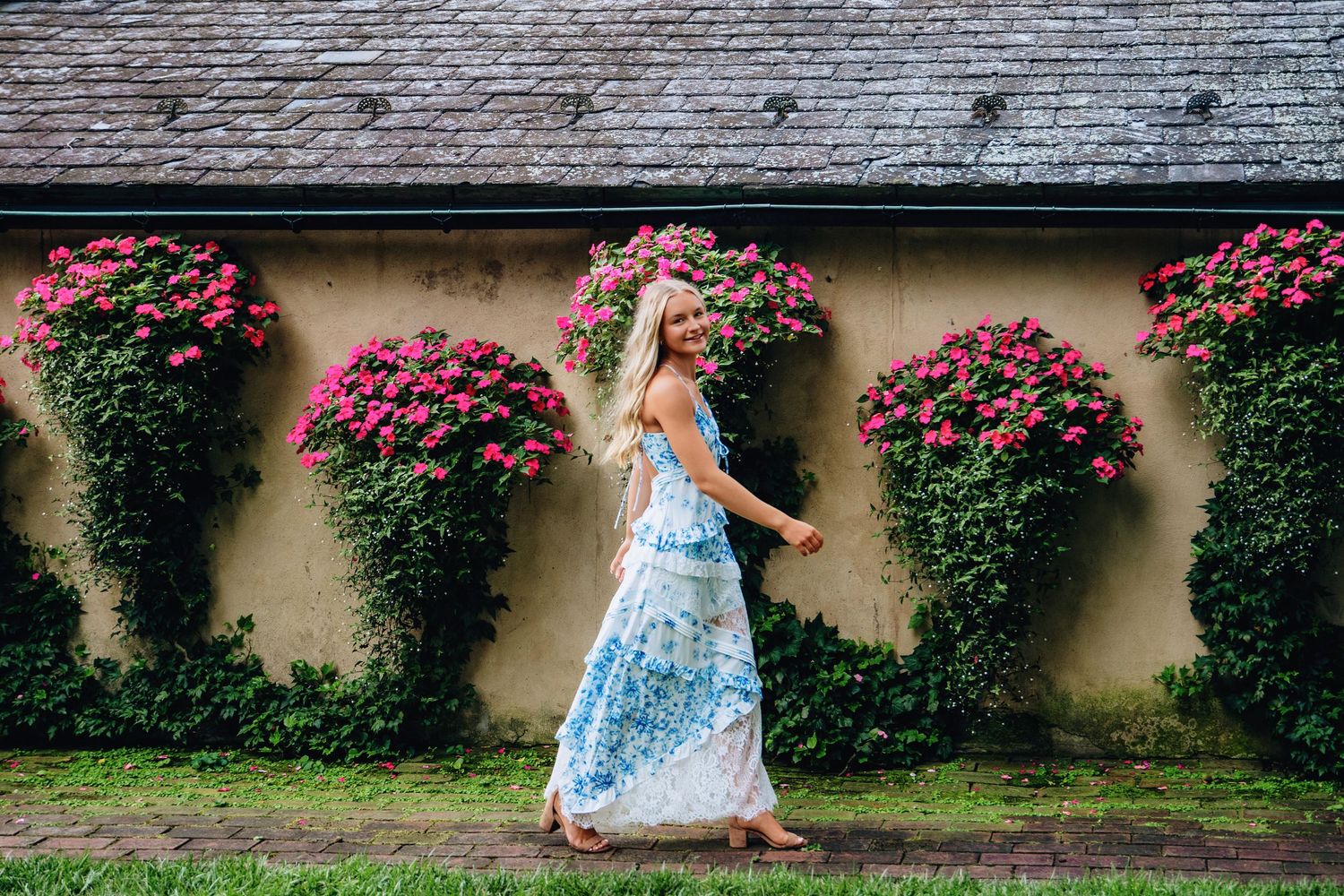 Woman in blue floral maxi dress walking beside a wall adorned with lush green ivy and vibrant pink flowers.