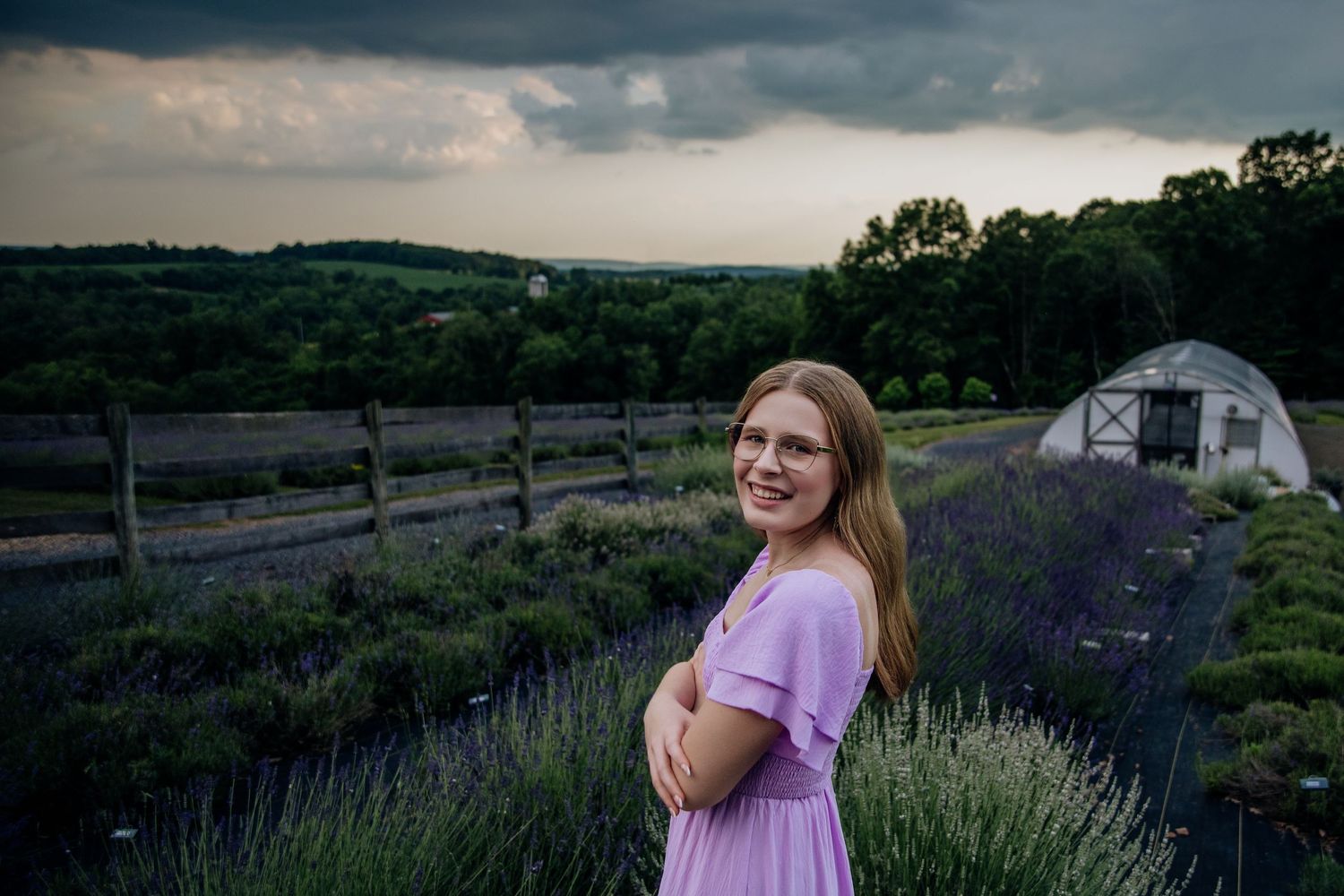 Young woman in lavender dress smiling in a lavender field with stormy skies and rolling hills.