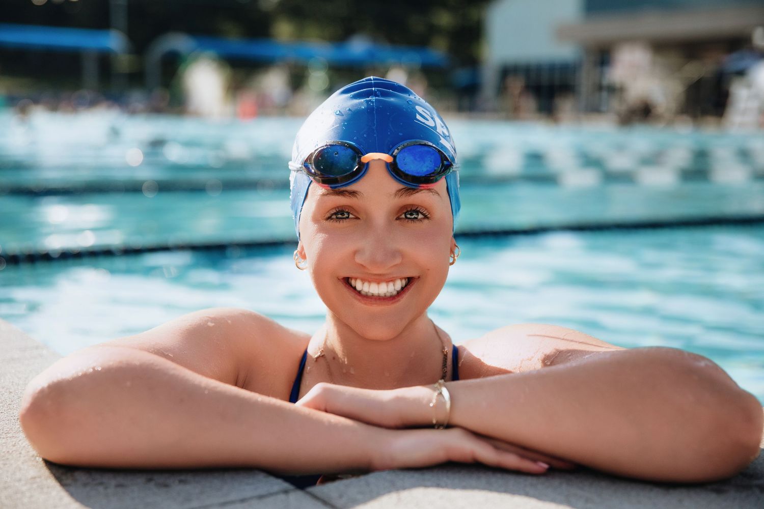 Smiling female swimmer in blue cap and goggles resting at pool edge on sunny day.