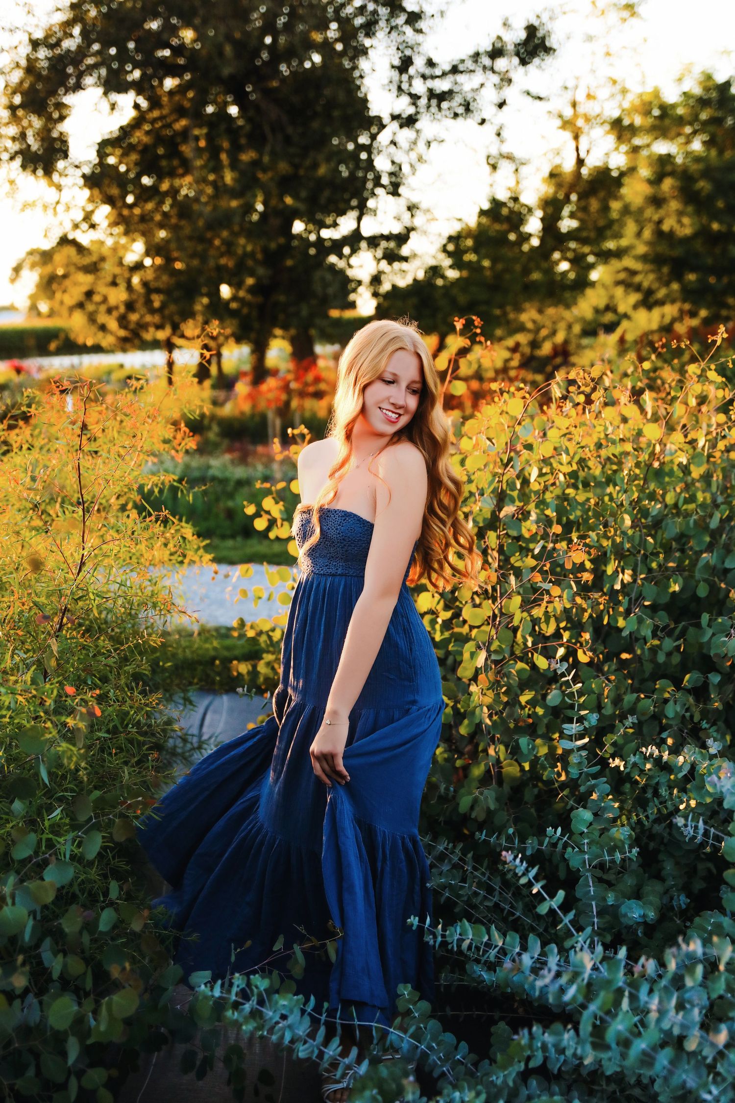 A woman in a strapless navy blue maxi dress poses gracefully among lush garden greenery in golden hour sunlight.