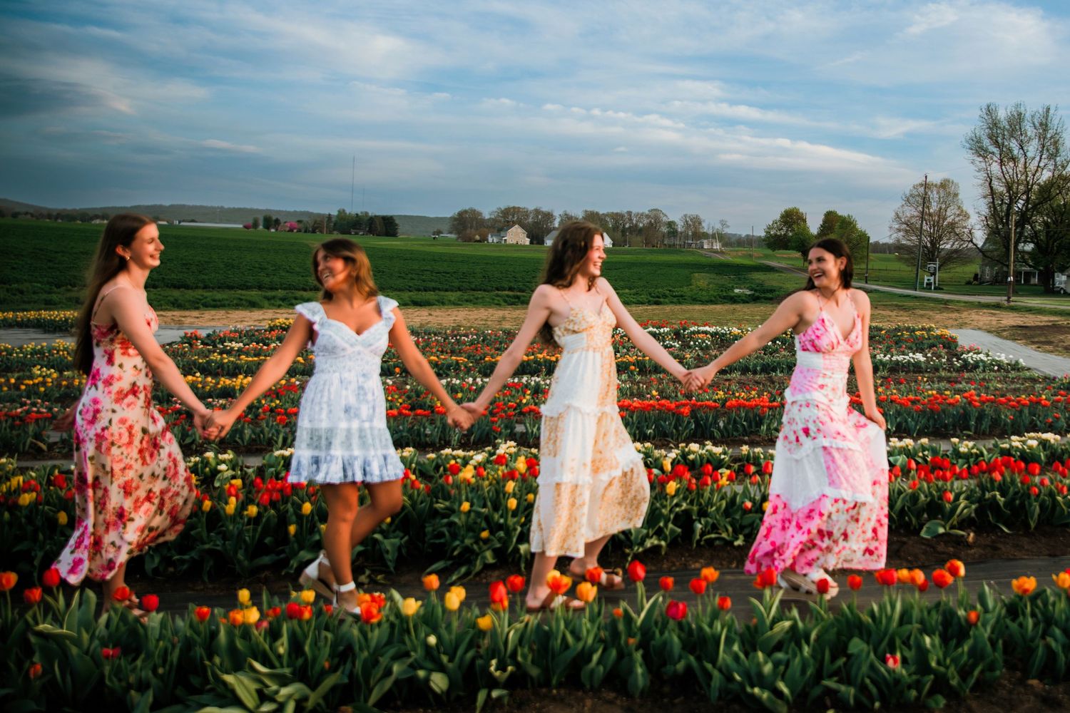 Four women in floral dresses hold hands walking through a colorful tulip field at golden hour.