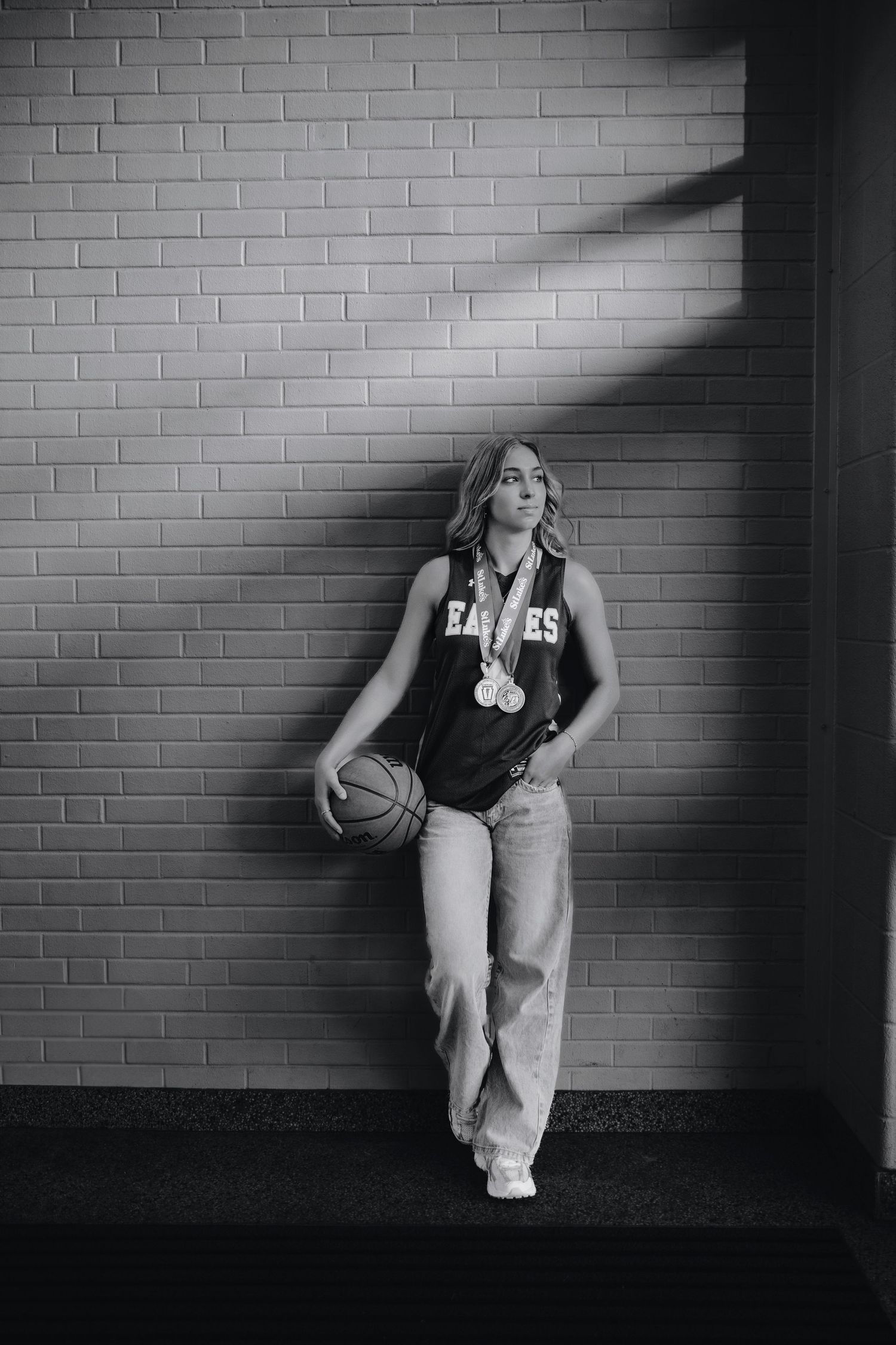 Black and white photo of a young female basketball player in Eagles jersey holding a ball, leaning against a brick wall.