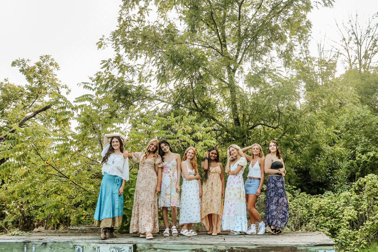 Group of eight young women in floral and pastel maxi dresses posing outdoors on a ledge with lush green trees behind them.