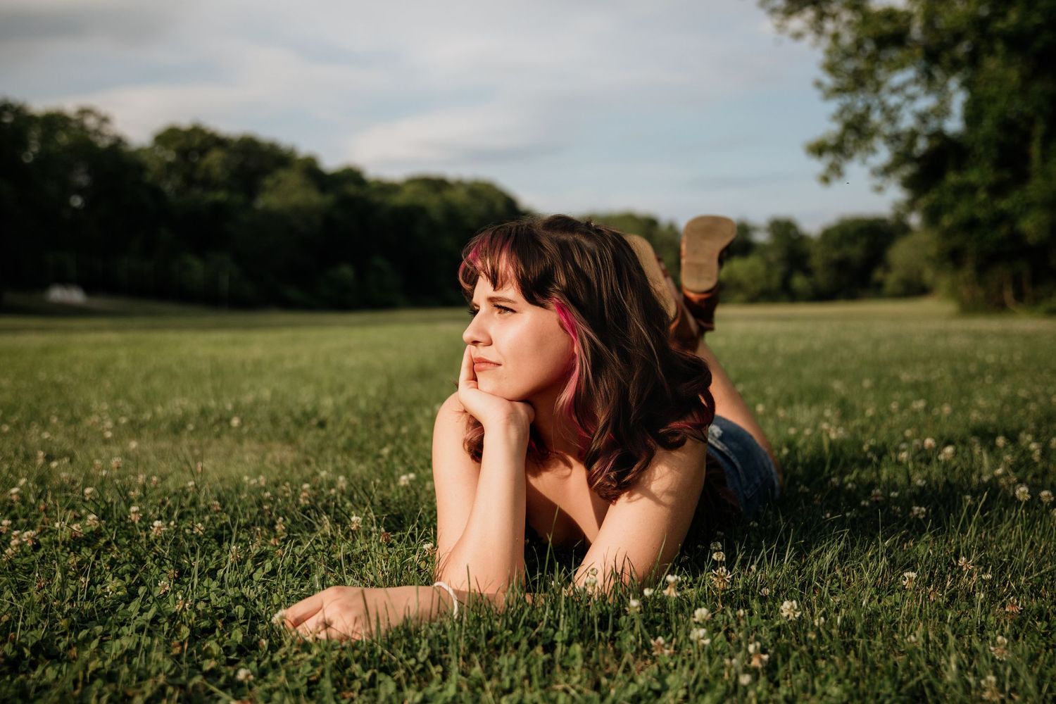Young woman with pink hair accessory lying on green grass in a sunny park, gazing thoughtfully into the distance.