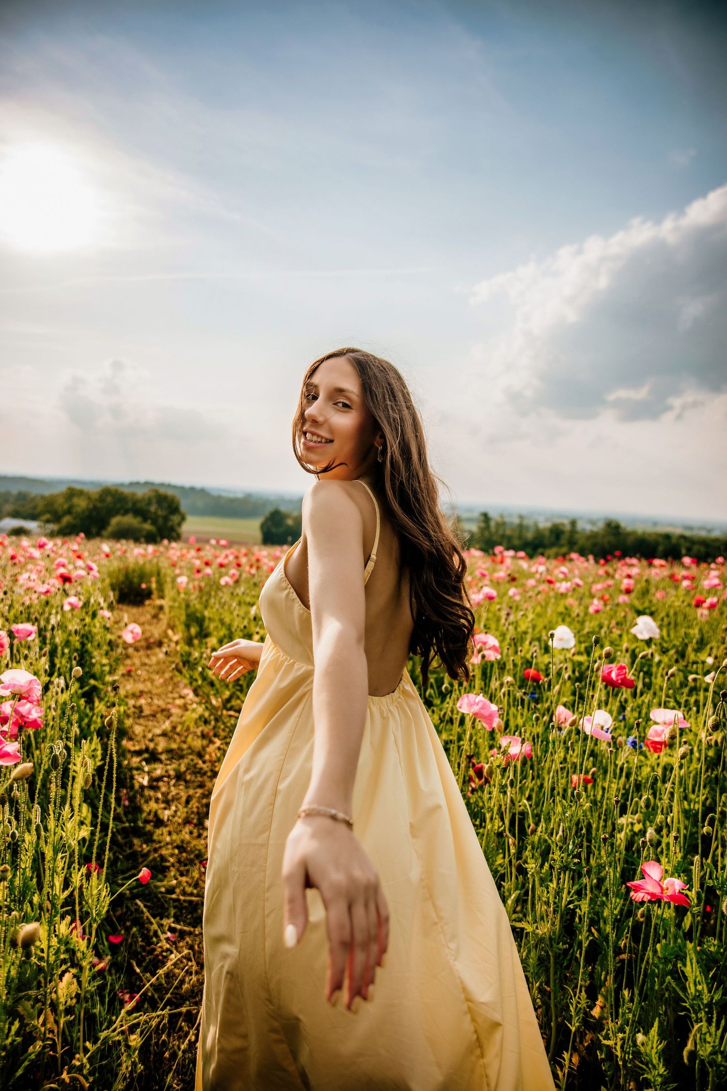 Young woman in yellow dress smiling in a vibrant pink poppy field under a sunny sky.