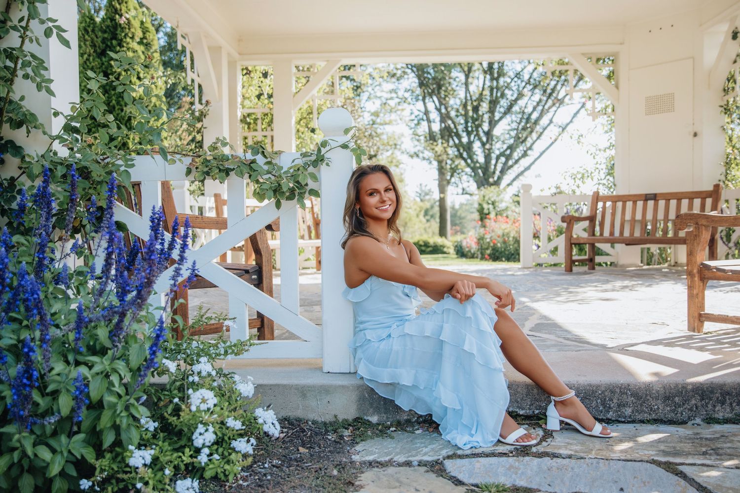 Young woman in light blue dress sitting on white steps surrounded by blue flowers and greenery in elegant garden setting.