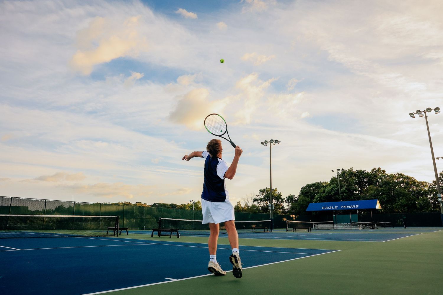 Tennis player serving on a blue outdoor court under a dramatic cloudy sky at dusk.