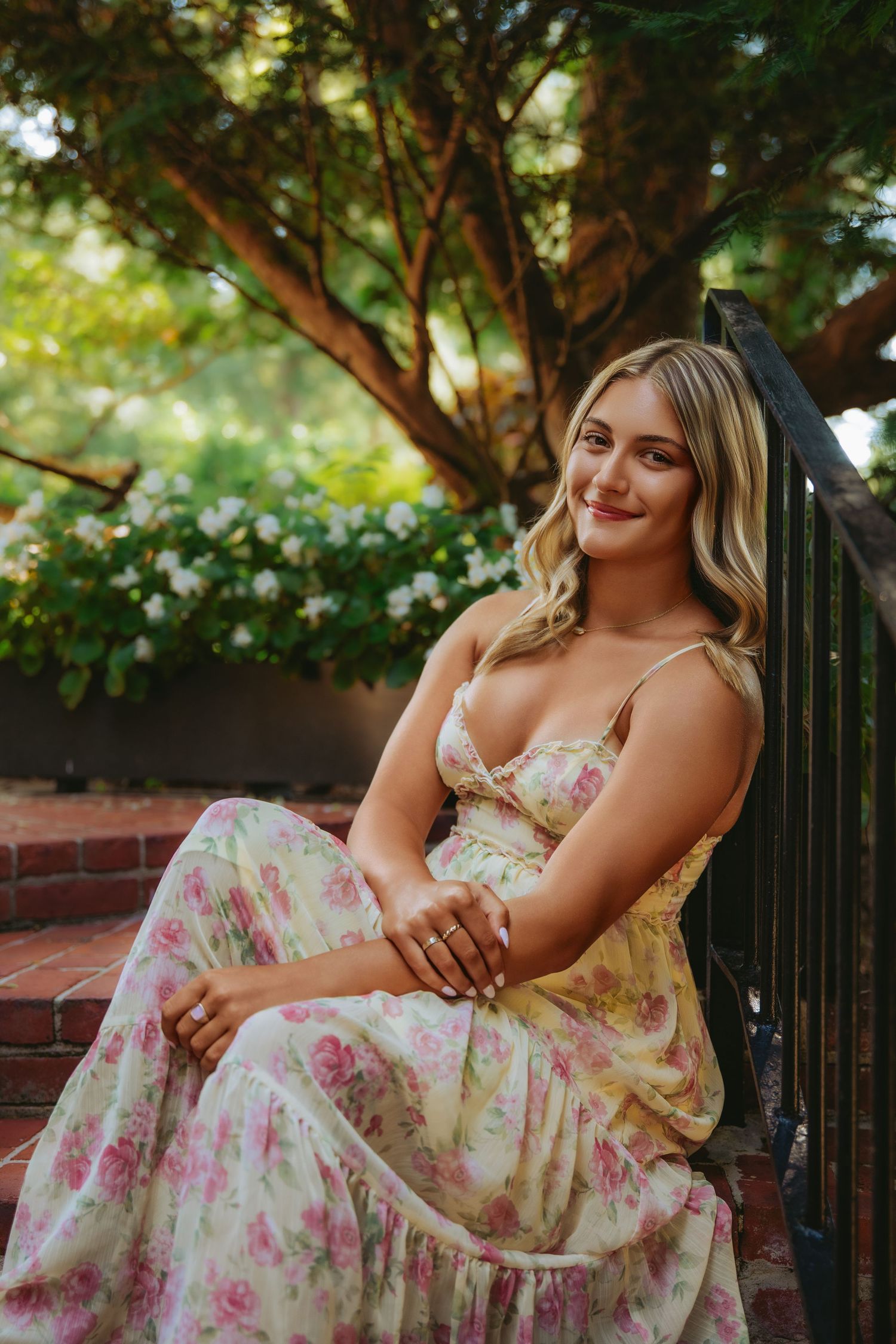 Young woman in floral maxi dress sitting on brick steps in a lush garden setting.