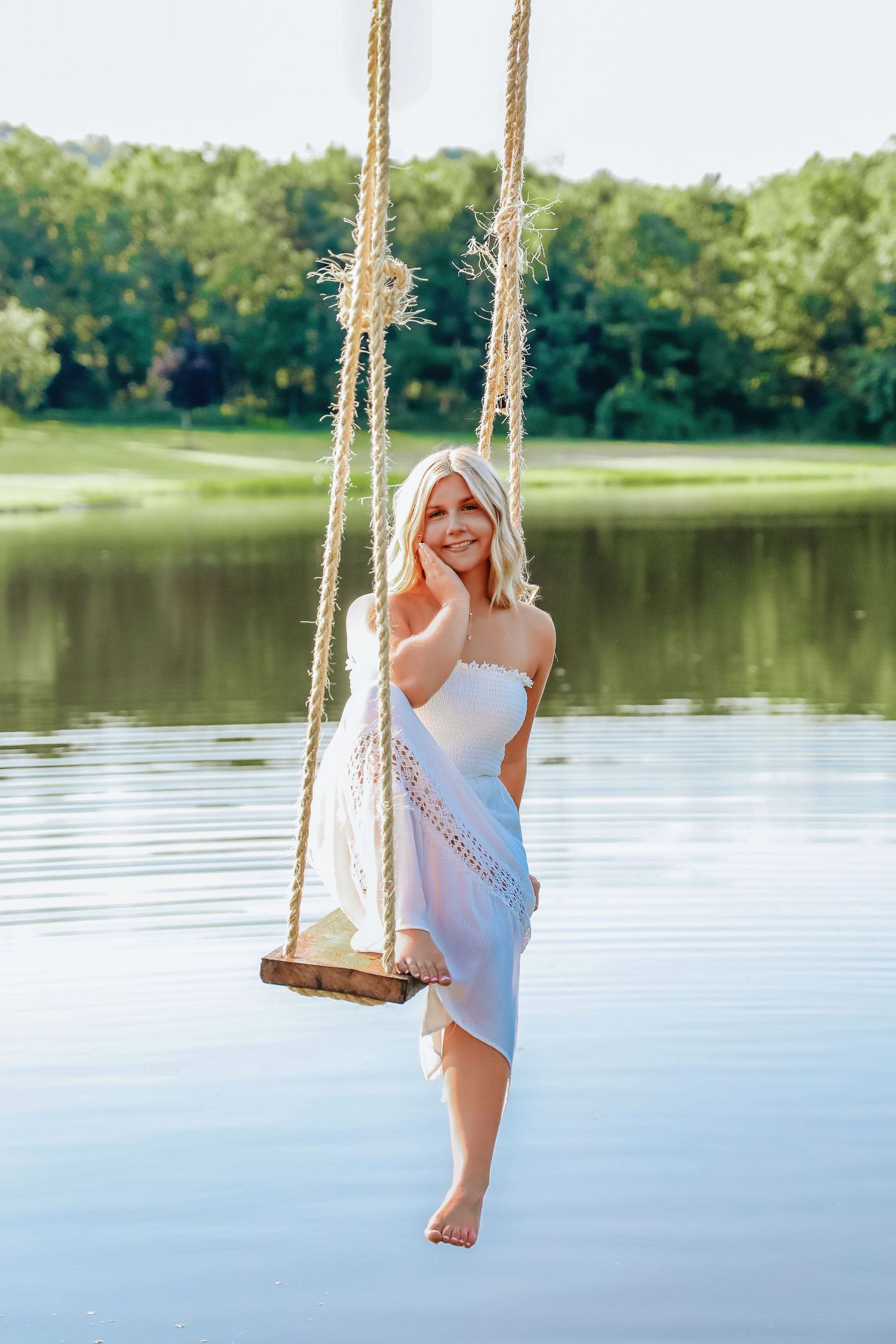 Smiling blonde woman in white dress sitting on rope swing over calm lake surrounded by lush green trees.