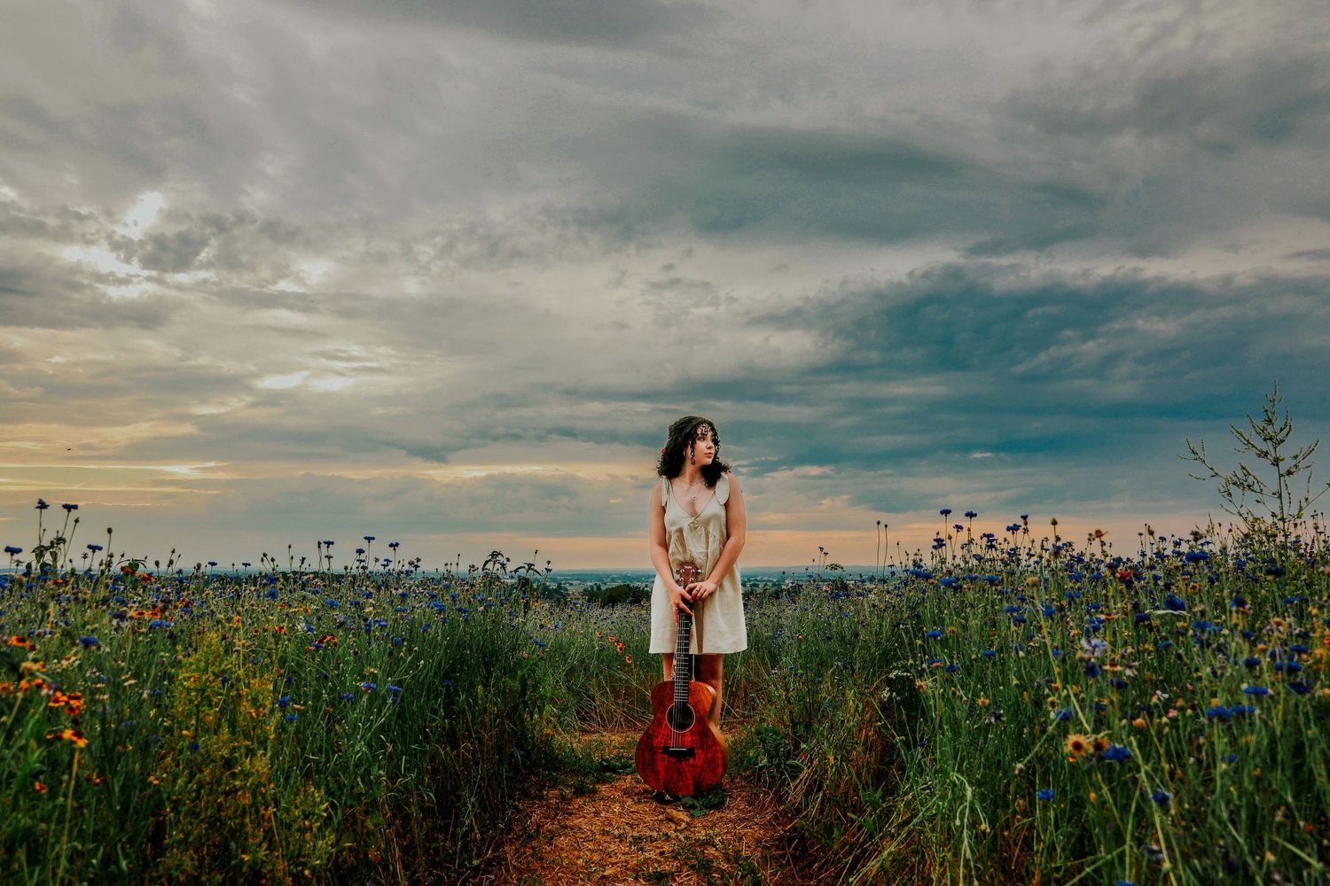 A young woman in a white dress stands in a wildflower field holding a red guitar under a dramatic cloudy sky.