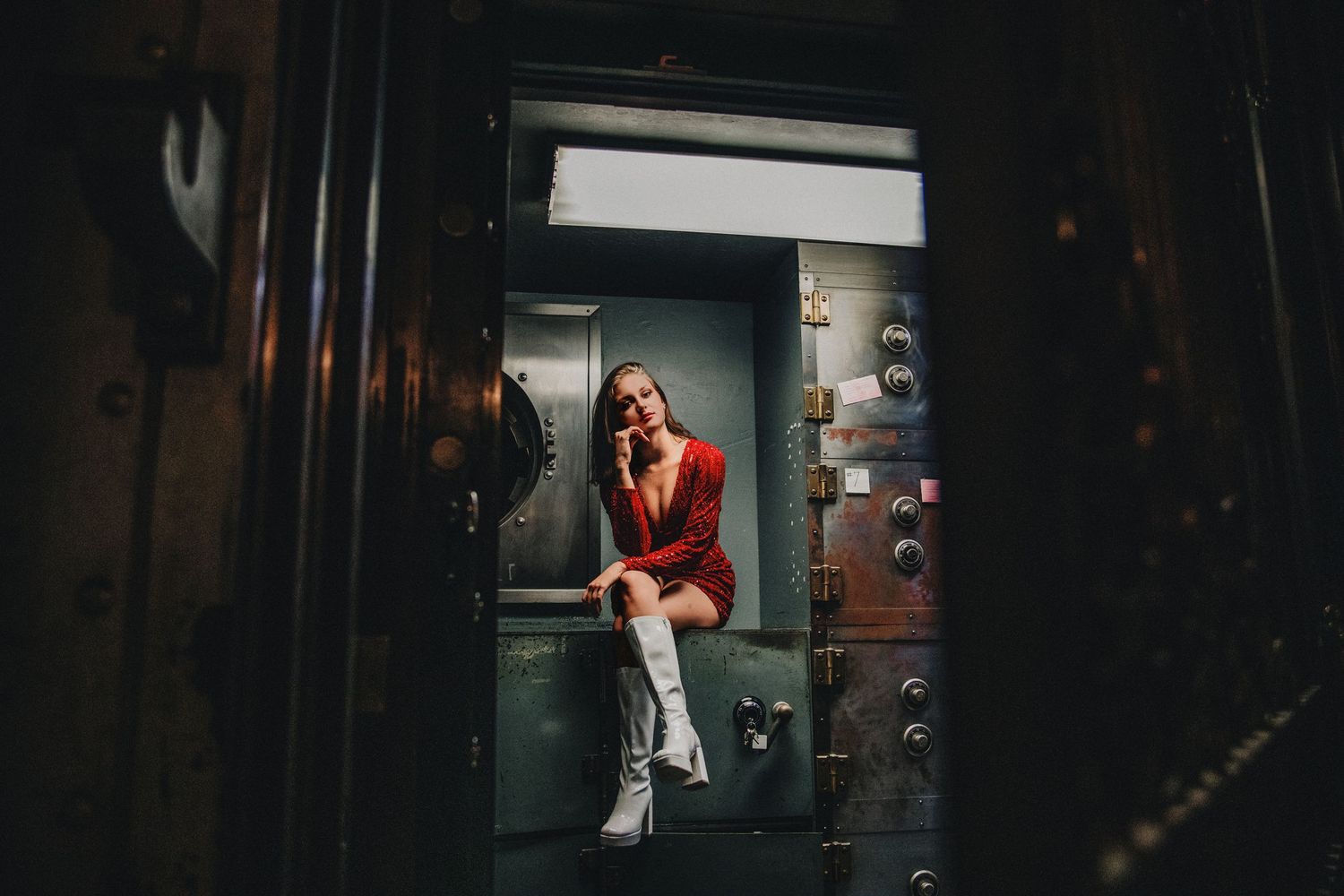 Woman in red dress and white boots poses inside a vintage bank vault door in dramatic moody lighting.