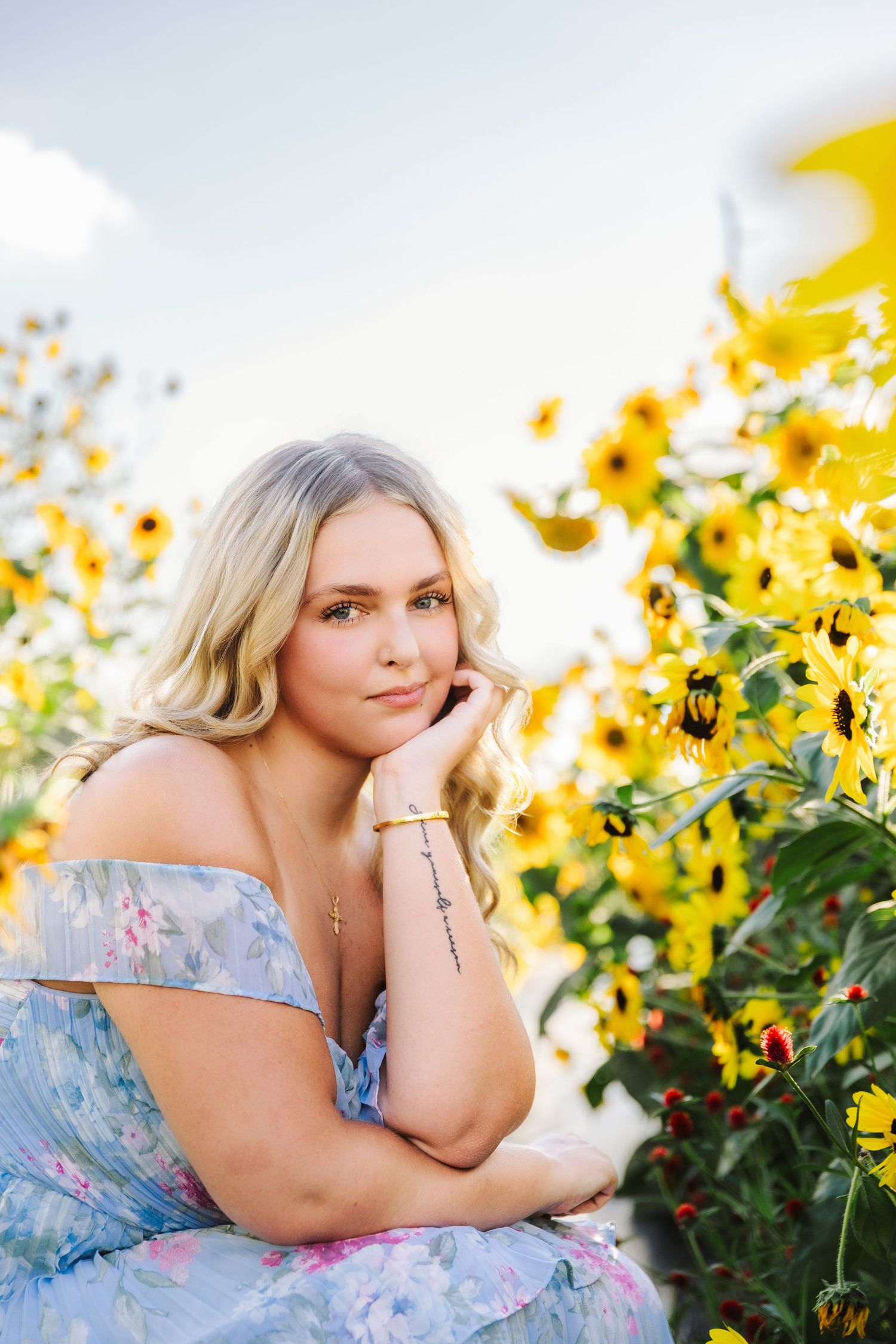 Blonde woman in blue floral off-shoulder dress posing among vibrant yellow sunflowers outdoors.