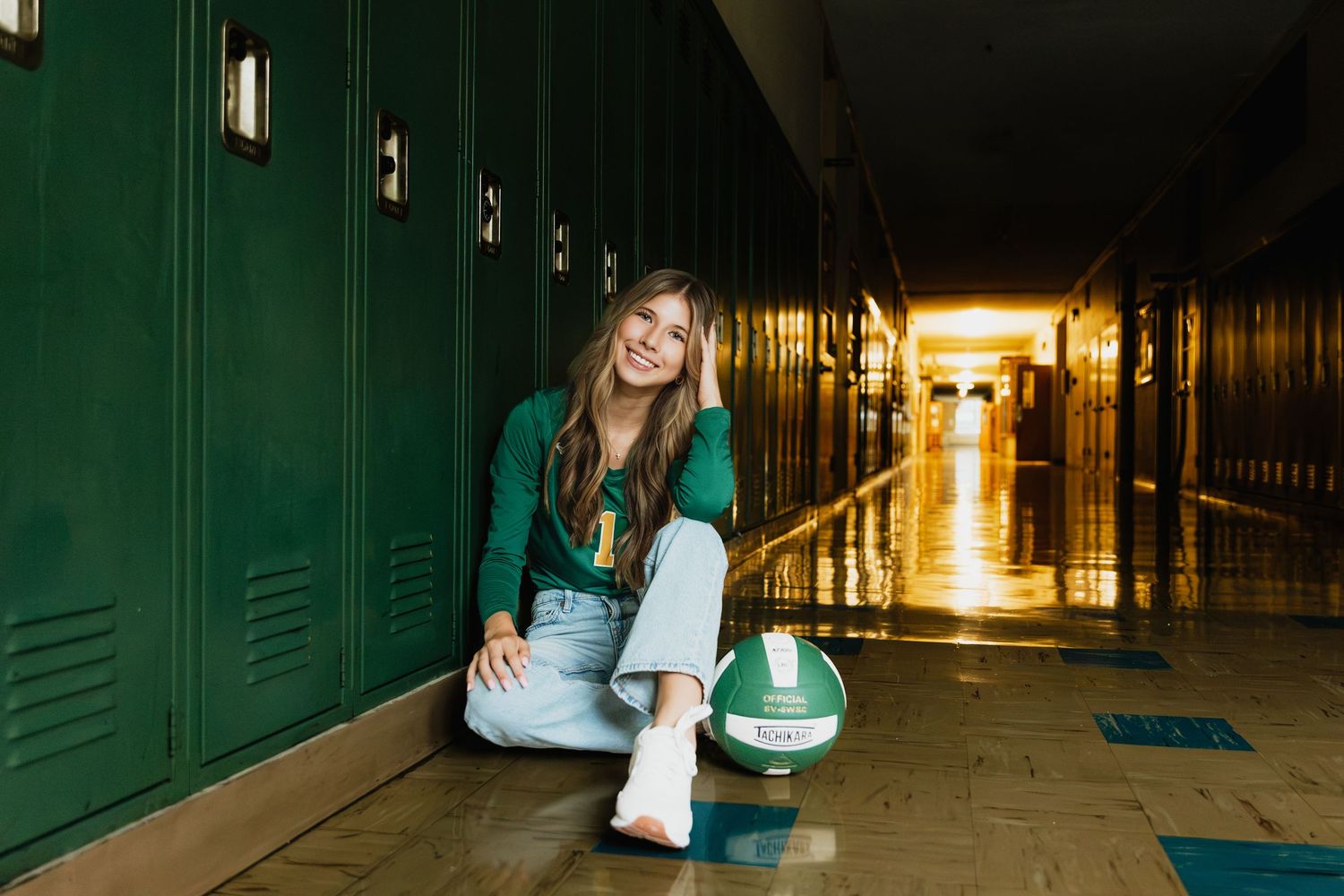 Teen girl in green sweater sits by school lockers with volleyball, smiling in a bright hallway for senior photos.
