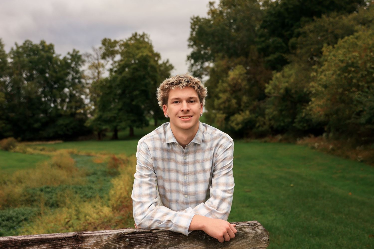 Young man in plaid shirt leaning on wooden fence in lush green park setting with trees in background.