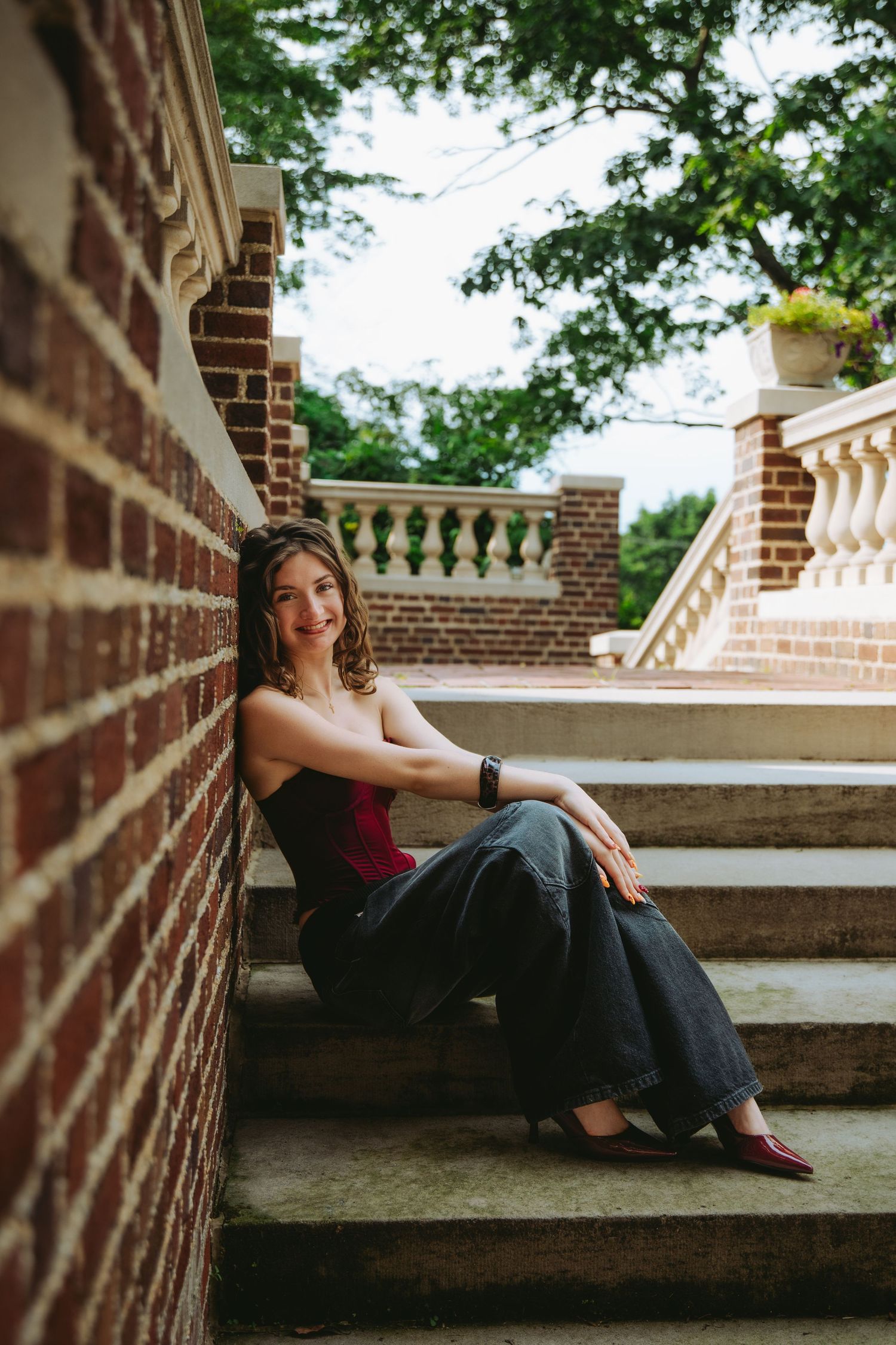 Young woman in red top and wide-leg jeans sitting on stone steps beside a brick wall, smiling at camera.