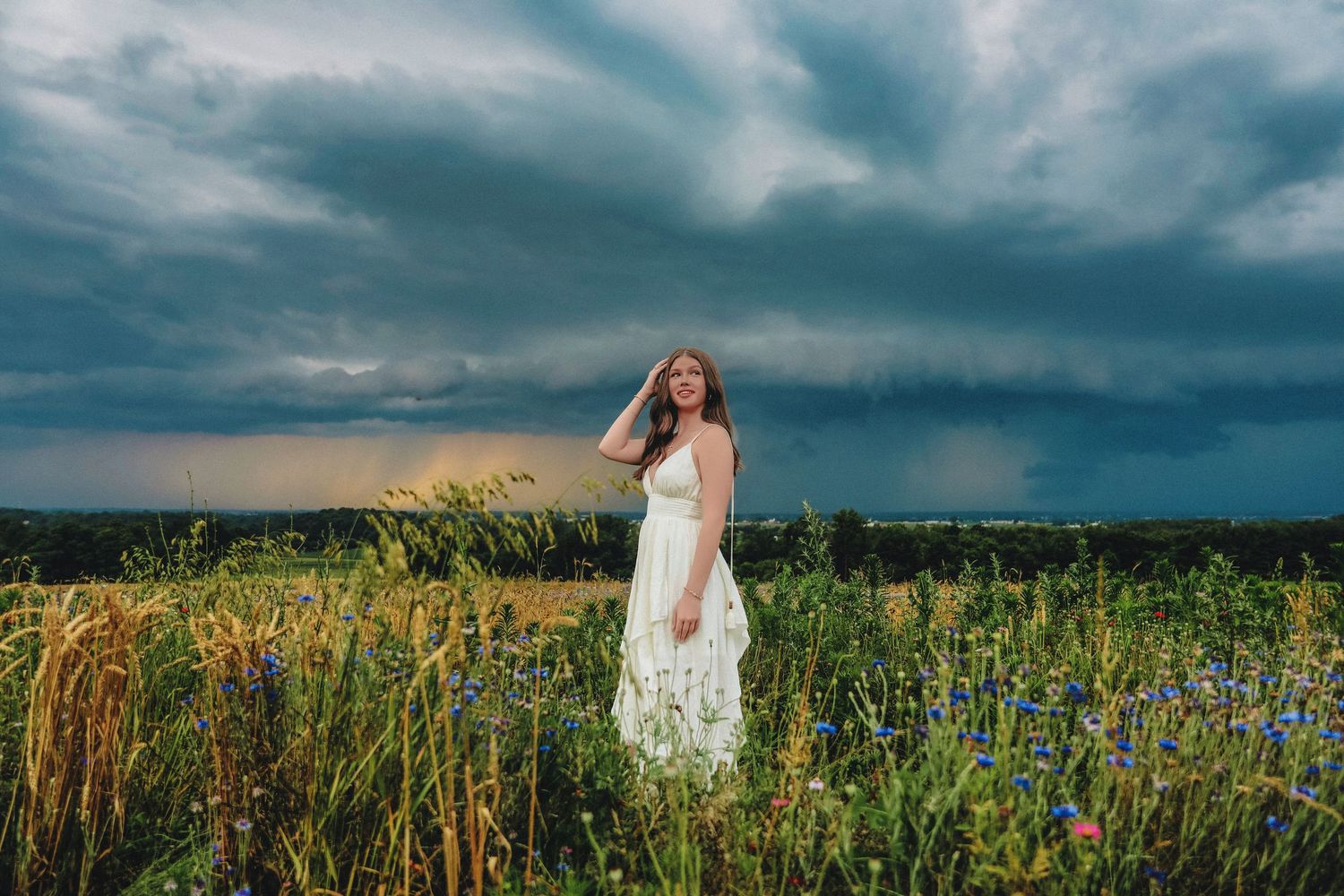 Young woman in white dress standing in wildflower field under dramatic stormy sky.