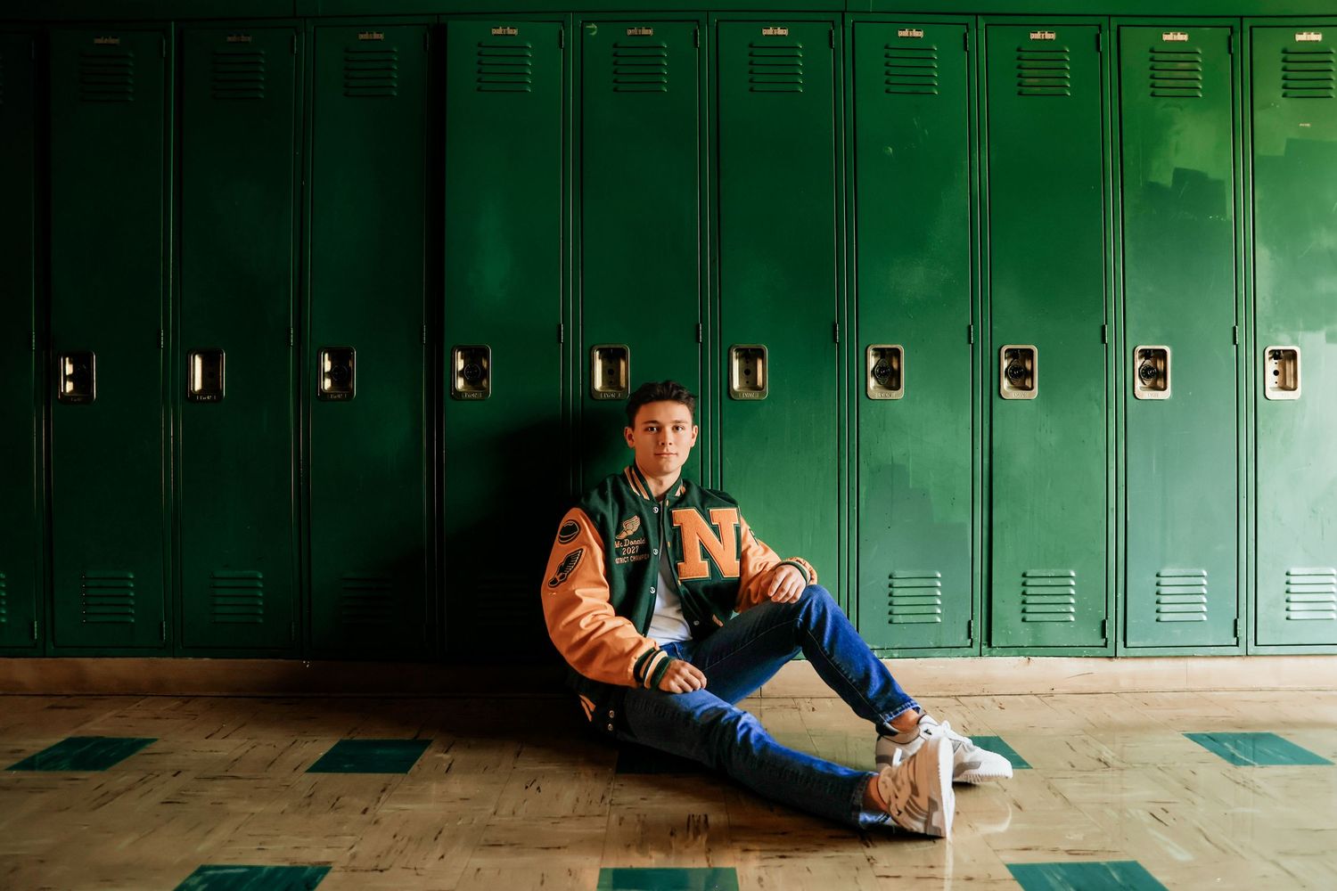 Teen boy in varsity jacket sits against green school lockers on hallway floor for senior portrait.