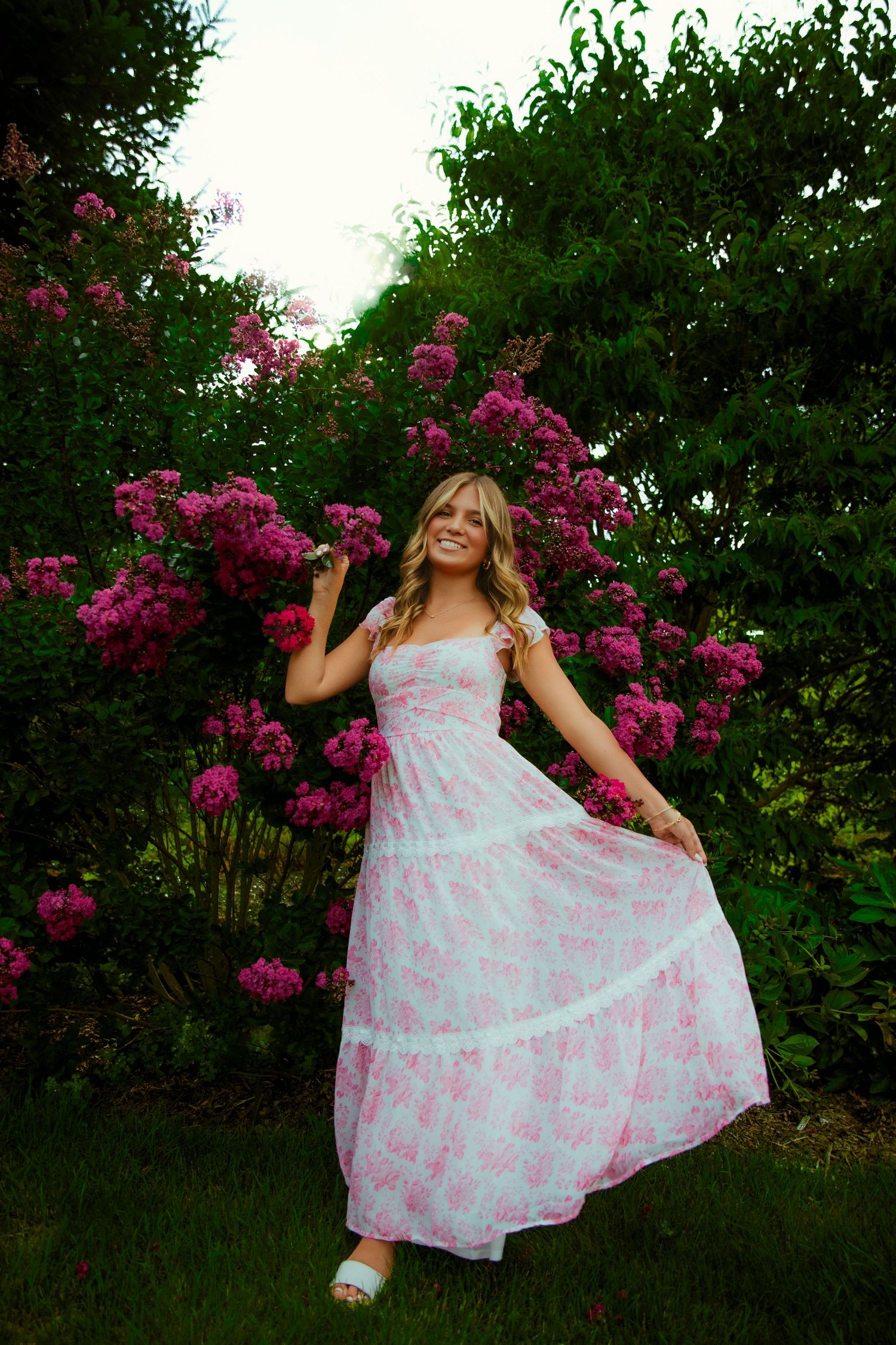 A smiling woman in a pink floral maxi dress poses gracefully among vibrant pink flowering bushes in a lush garden.